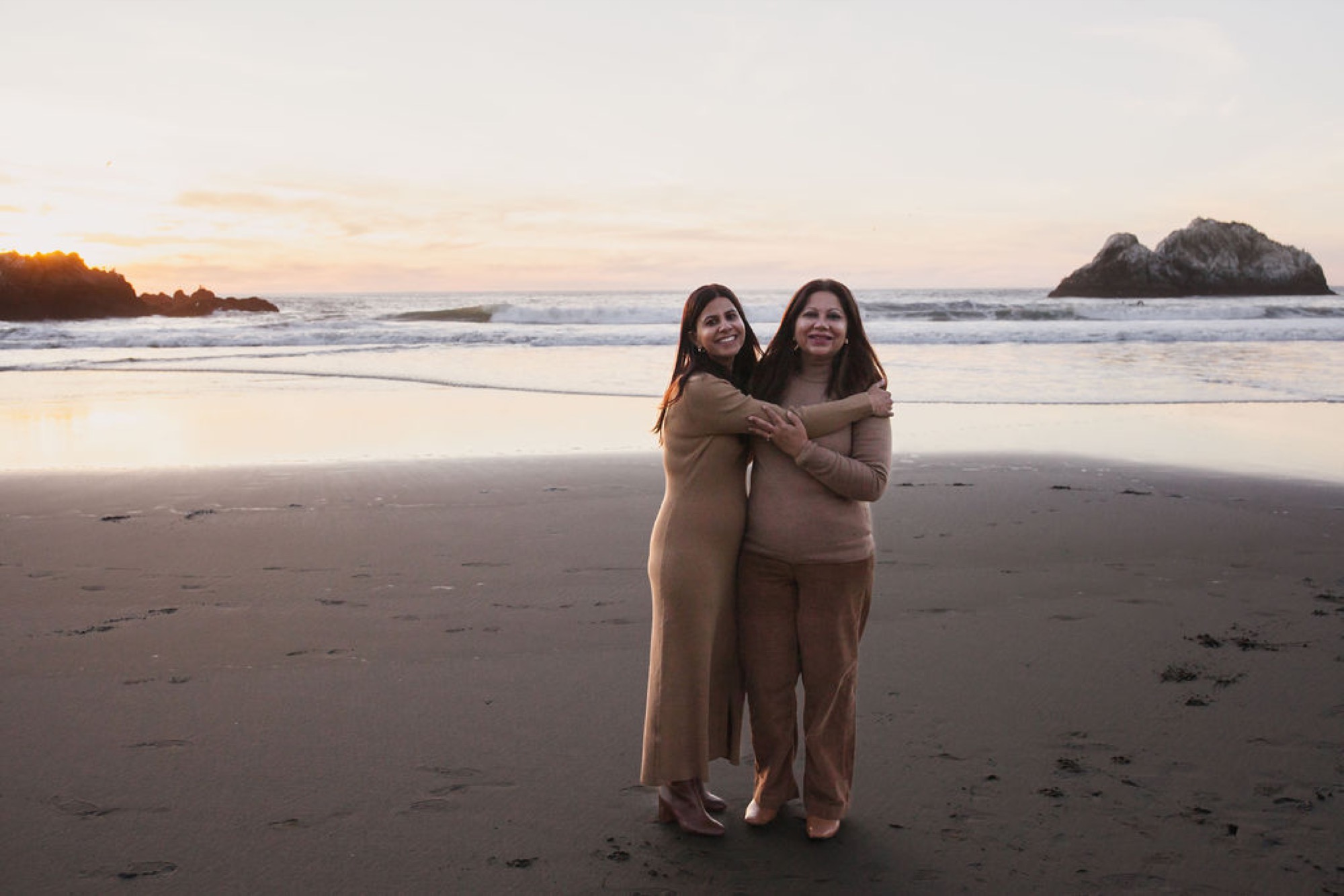 Two women embrace on a dark sand beach at sunset, with rocky islands and gentle waves framing the golden-hour moment