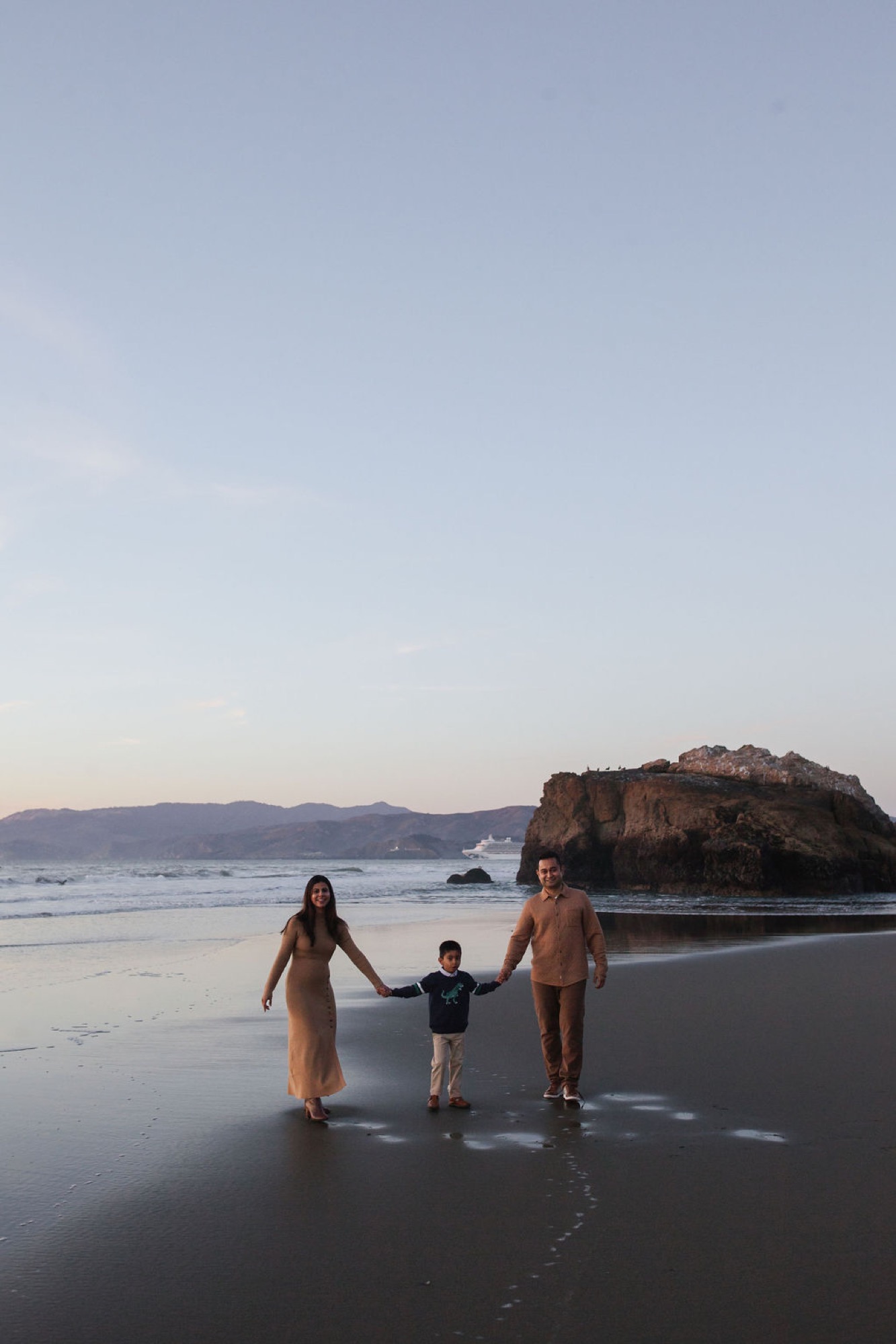 Family of three walks hand-in-hand along wet sand at sunset, with a rocky island and cruise ship visible across the bay
