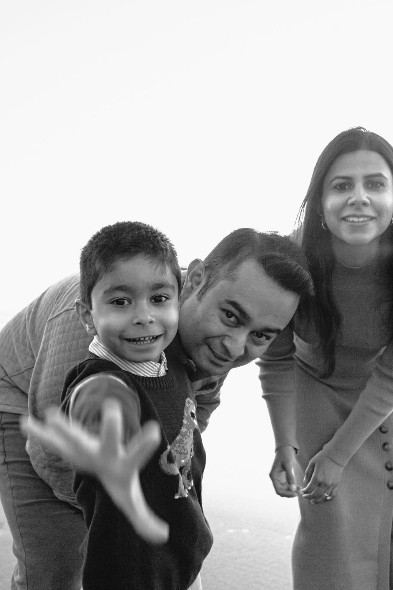 A young boy grins widely while pointing at the camera as his father leans in playfully, with his mother smiling warmly behind them against a bright overcast sky