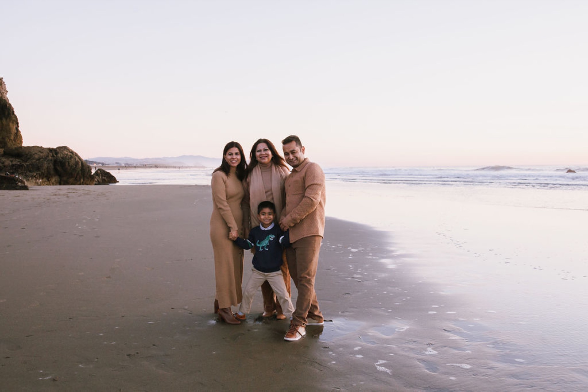 Four family members stand hand-in-hand on wet sand at sunset, their earth-toned clothing complementing the golden-hour light over the Pacific Ocean