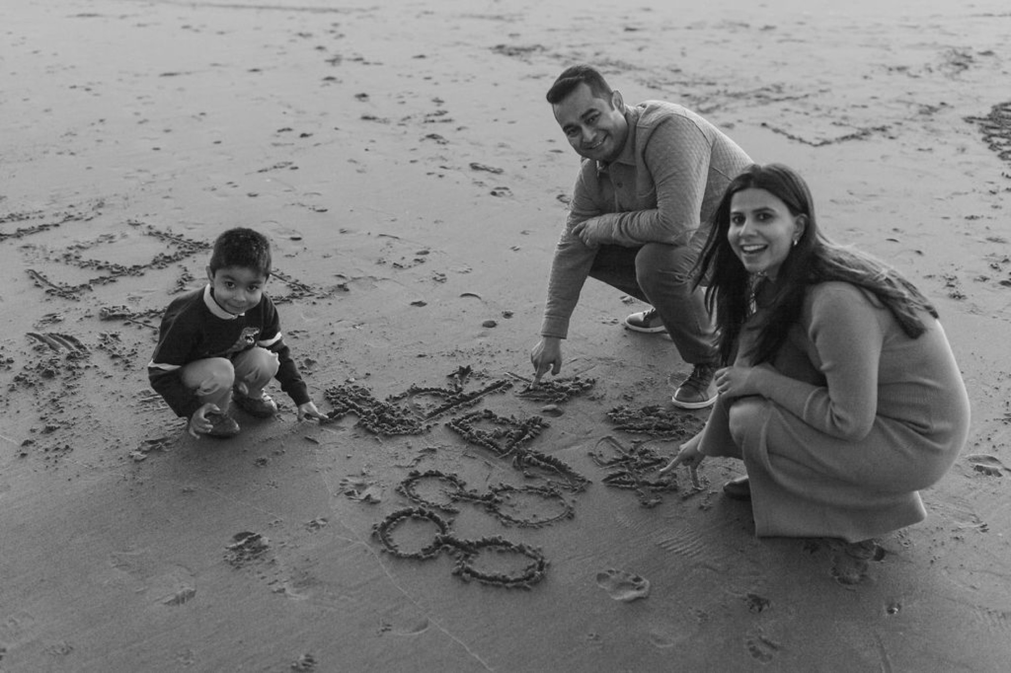 Family of four crouches together on wet sand, examining their names traced in the beach, genuine smiles and engaged postures
