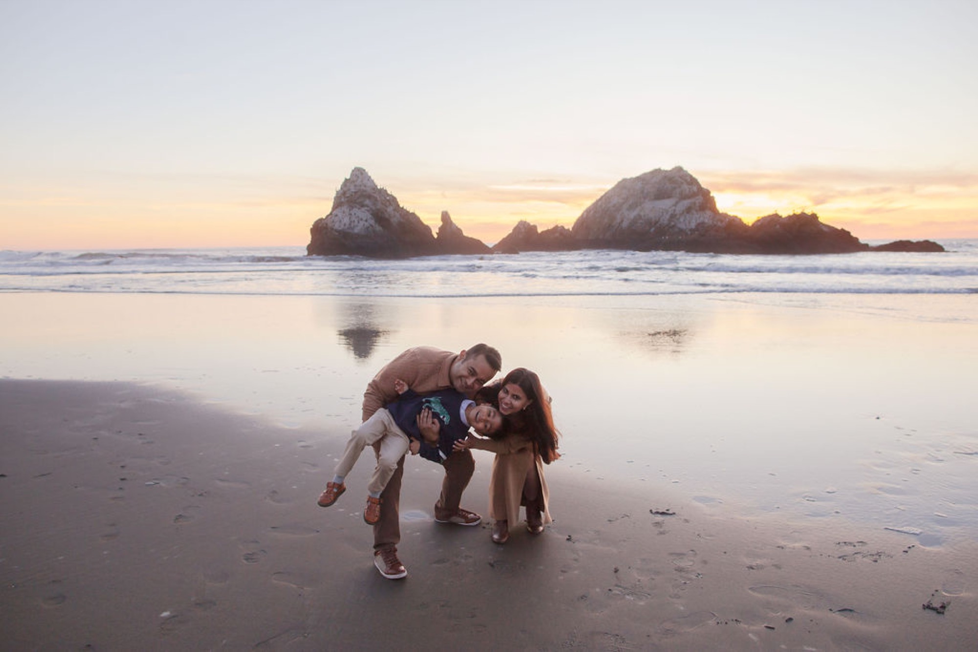 Father carries young child while mother and older daughter huddle close, silhouetted against golden sunset over dramatic sea stacks