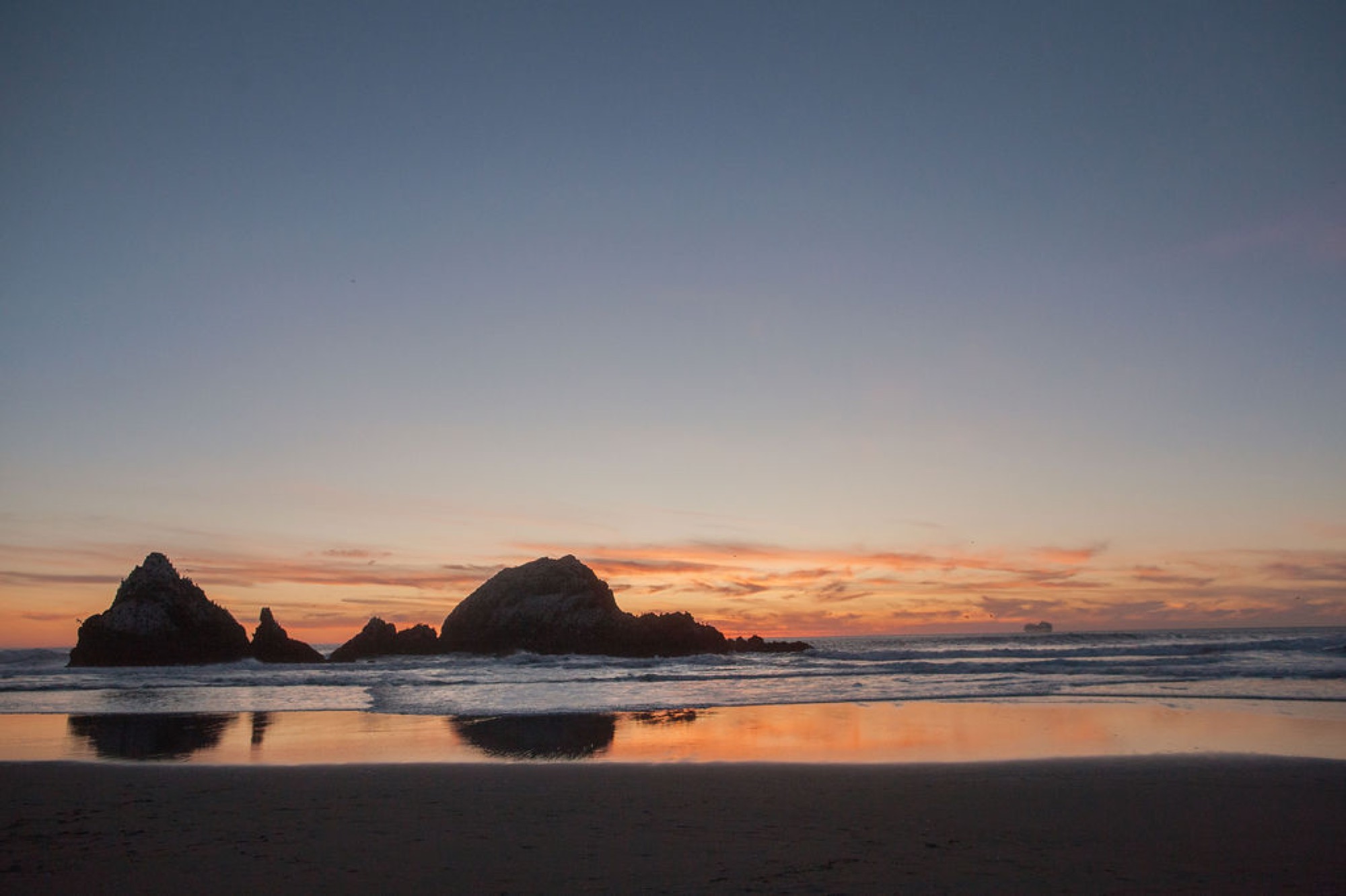 Silhouetted sea stacks glow beneath a golden sunset while their reflections mirror across the wet beach sand