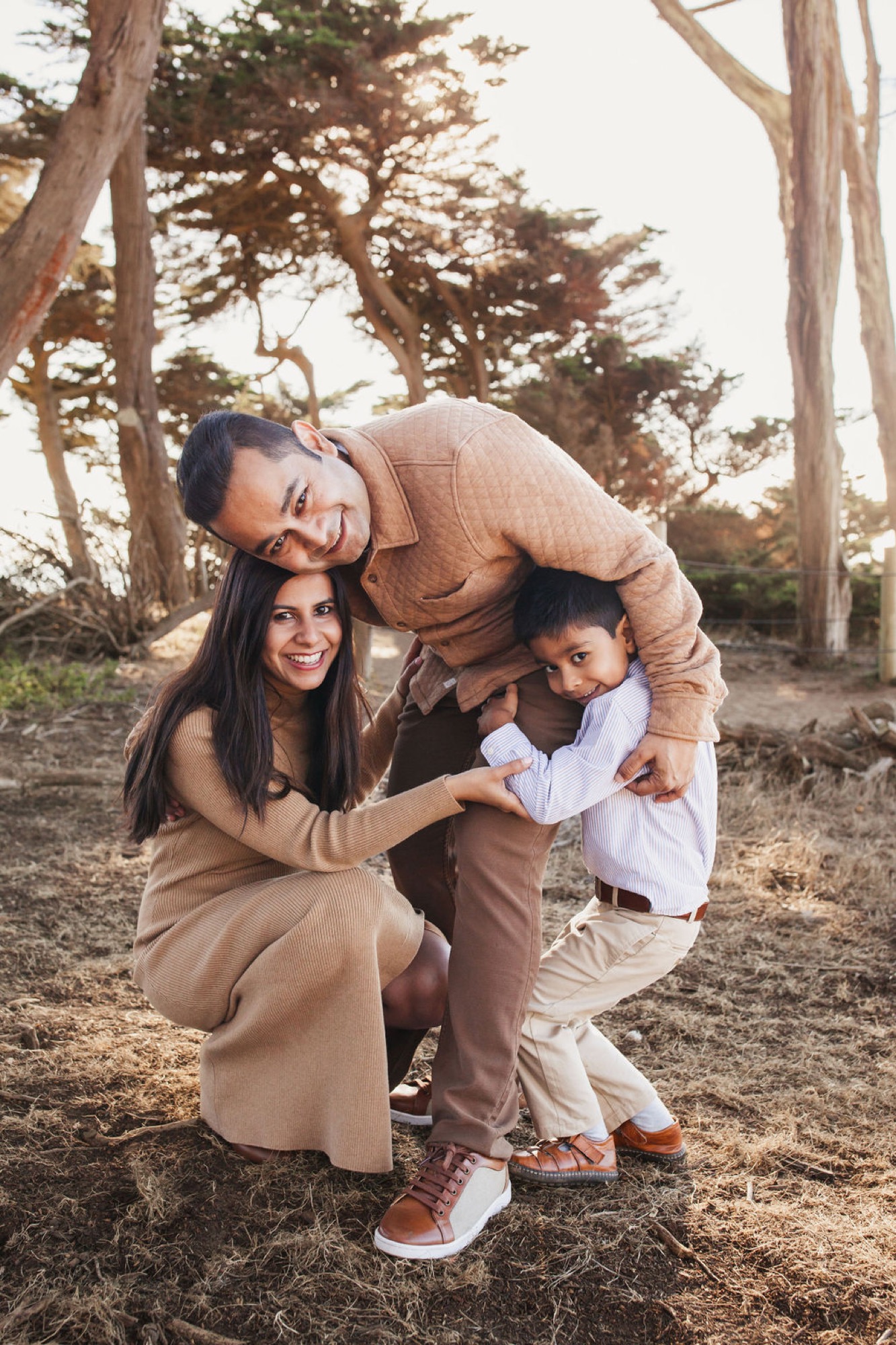 A father leans over to embrace his young son while the mother kneels beside them, all three smiling warmly beneath towering cypress trees bathed in golden afternoon light