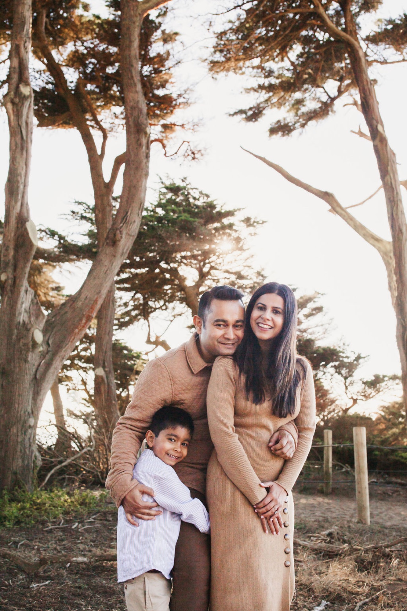 A father embraces his young son while the mother stands beside them, all smiling warmly beneath towering Monterey cypress trees in golden afternoon light