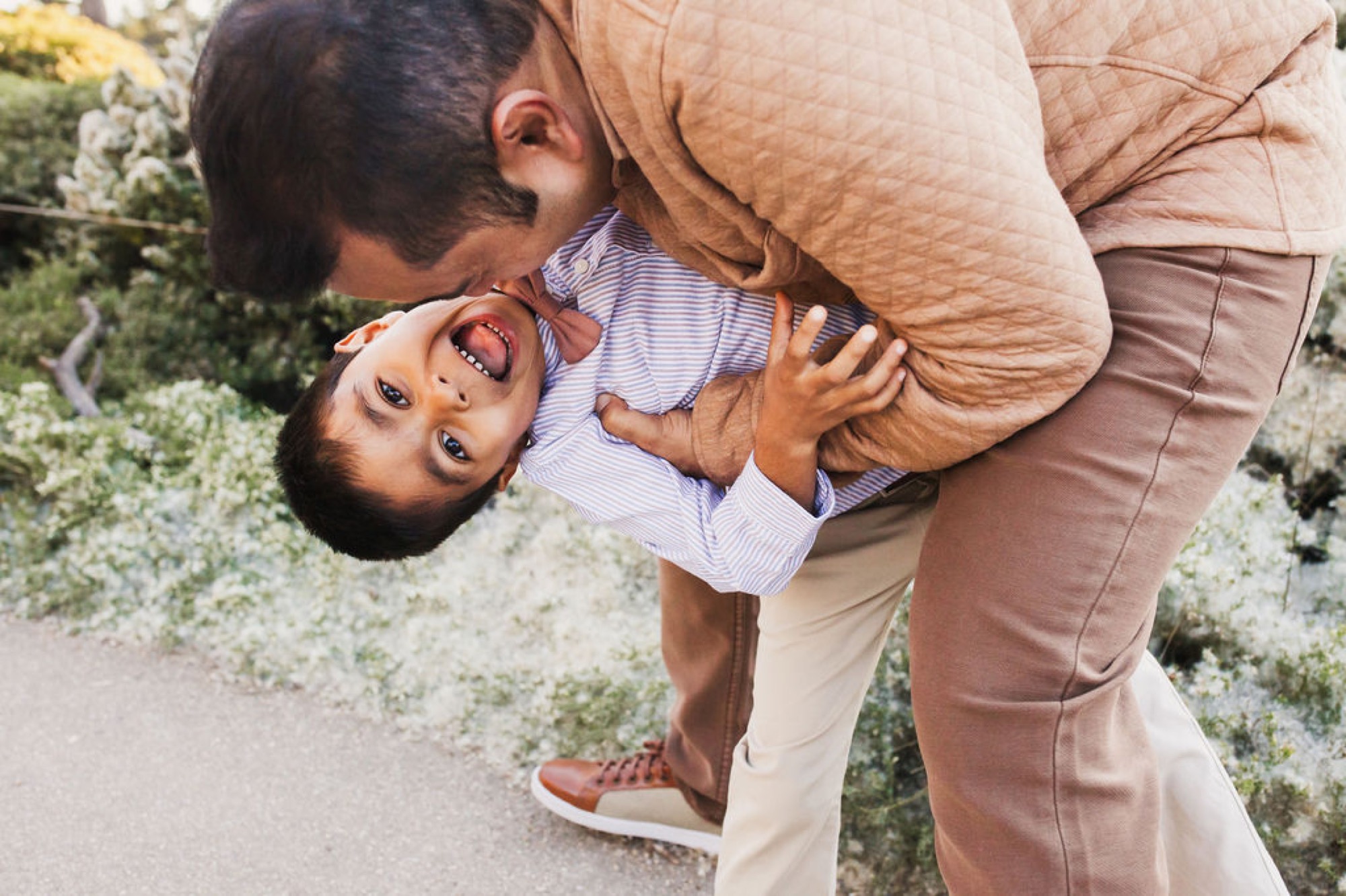 A father holds his young son upside down on a coastal path, the boy's delighted expression and open mouth capturing pure joy and playful affection