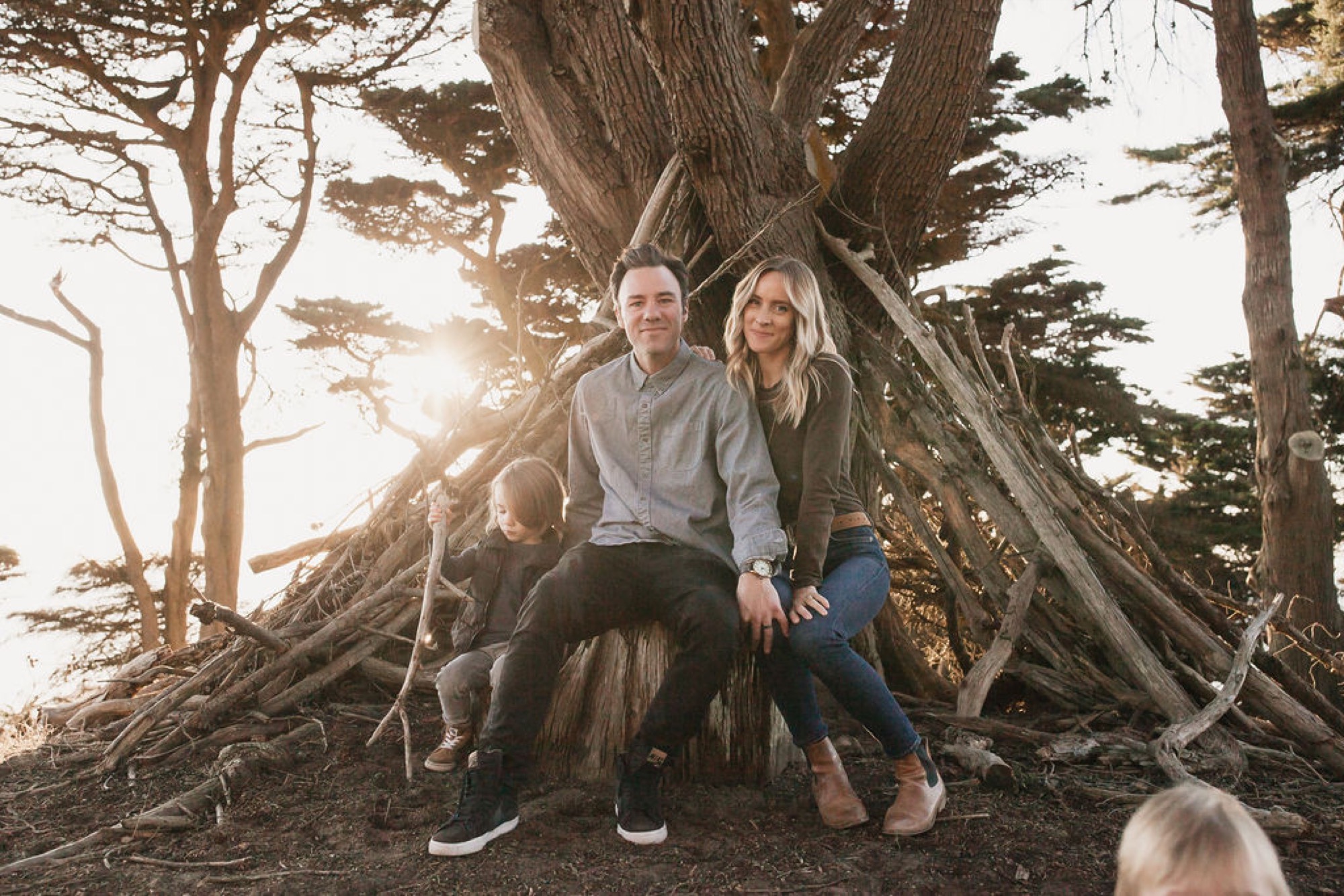 A couple sits shoulder-to-shoulder on massive gnarled tree roots at sunset, golden light filtering through windswept cypress branches behind them
