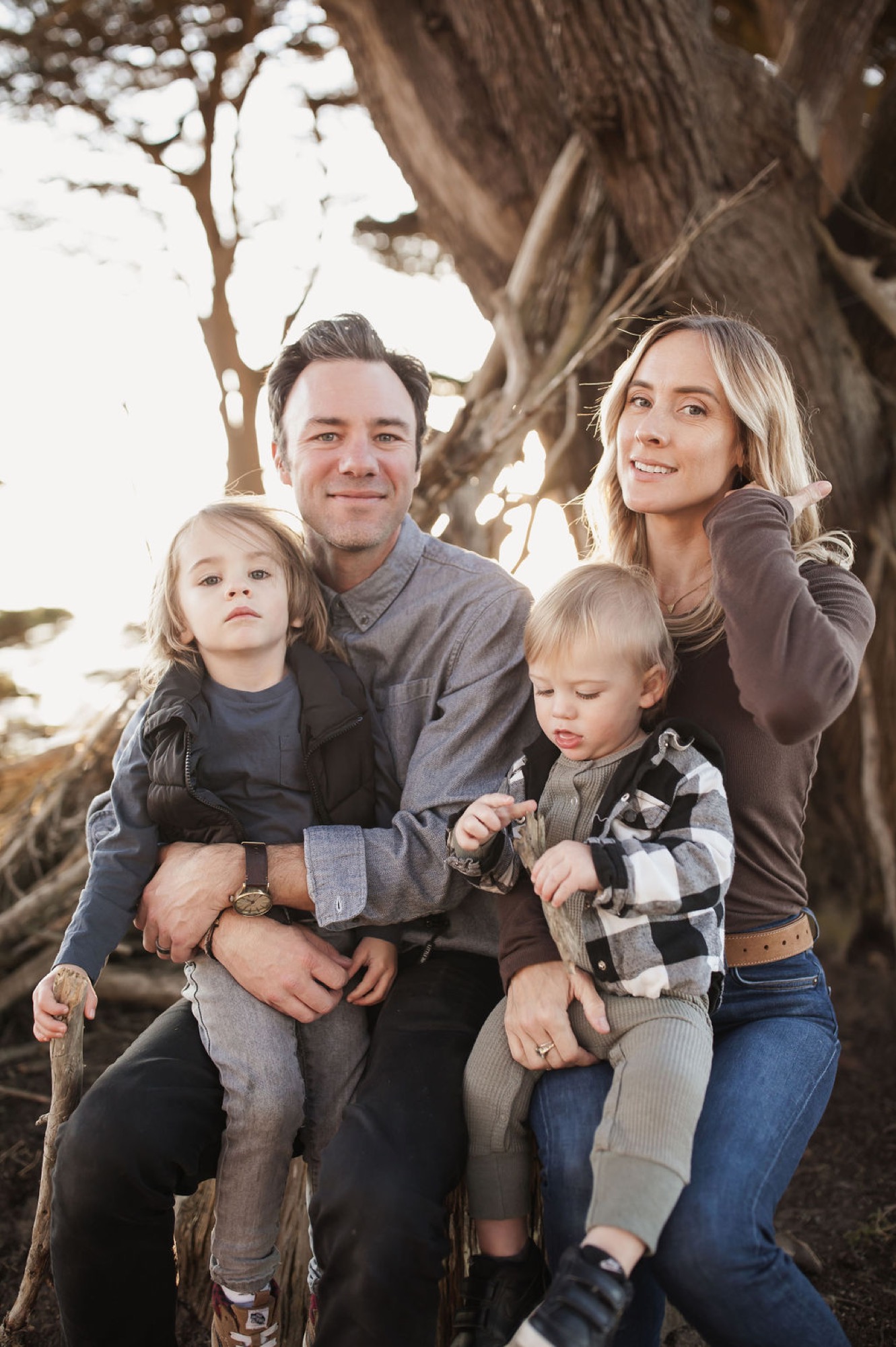 Family of four nestled beneath a gnarled tree trunk, sharing genuine smiles in golden afternoon light at the coast