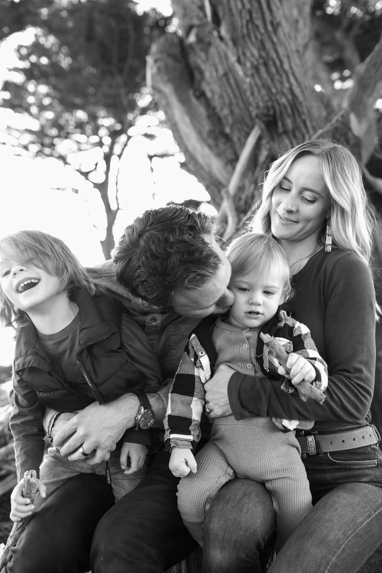 A mother and three children laugh together beneath a gnarled tree, their genuine joy captured in black and white against dappled natural light