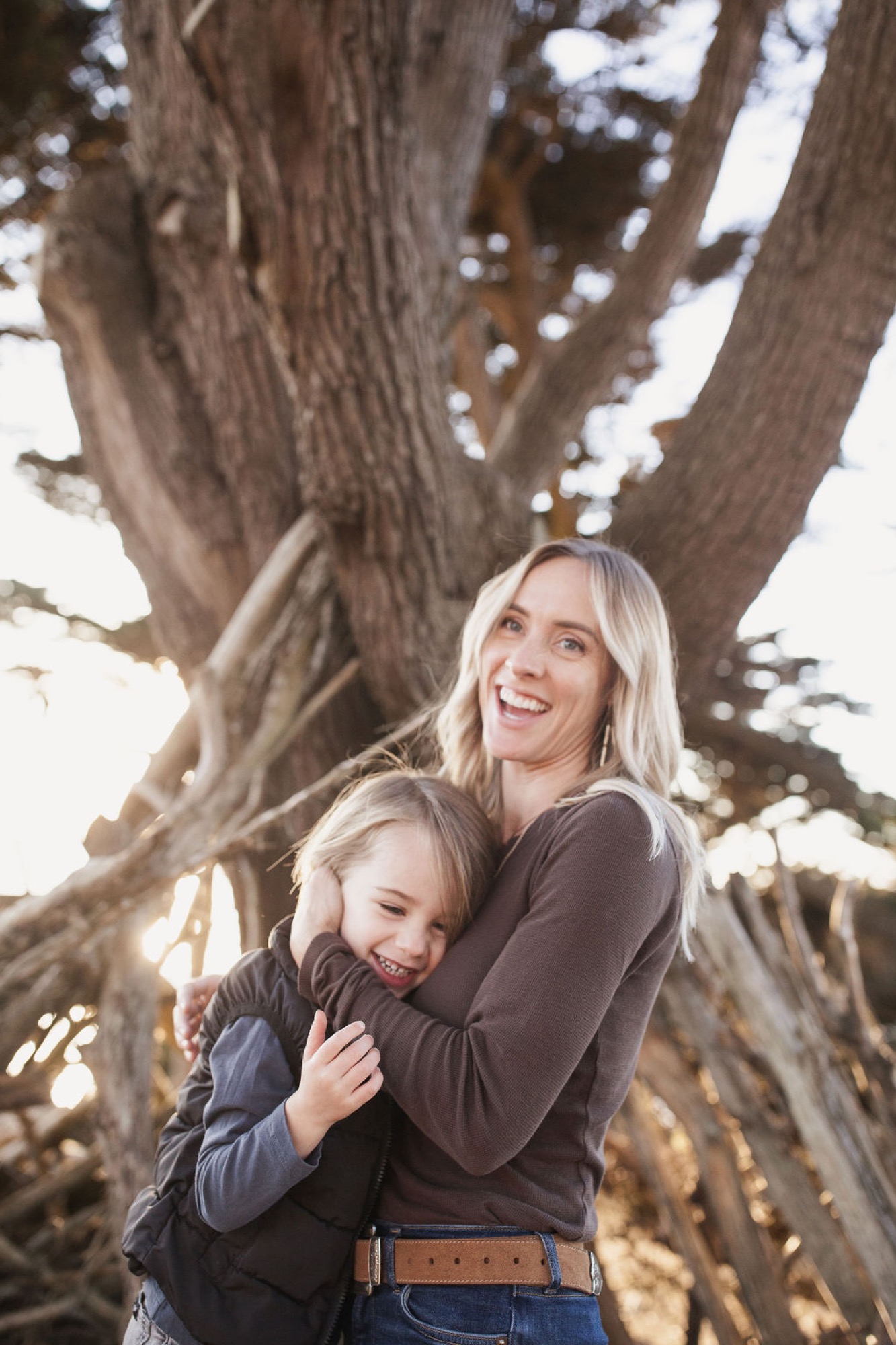 A blonde mother and young daughter embrace beneath a massive gnarled tree, both smiling warmly as dappled sunlight filters through the branches above