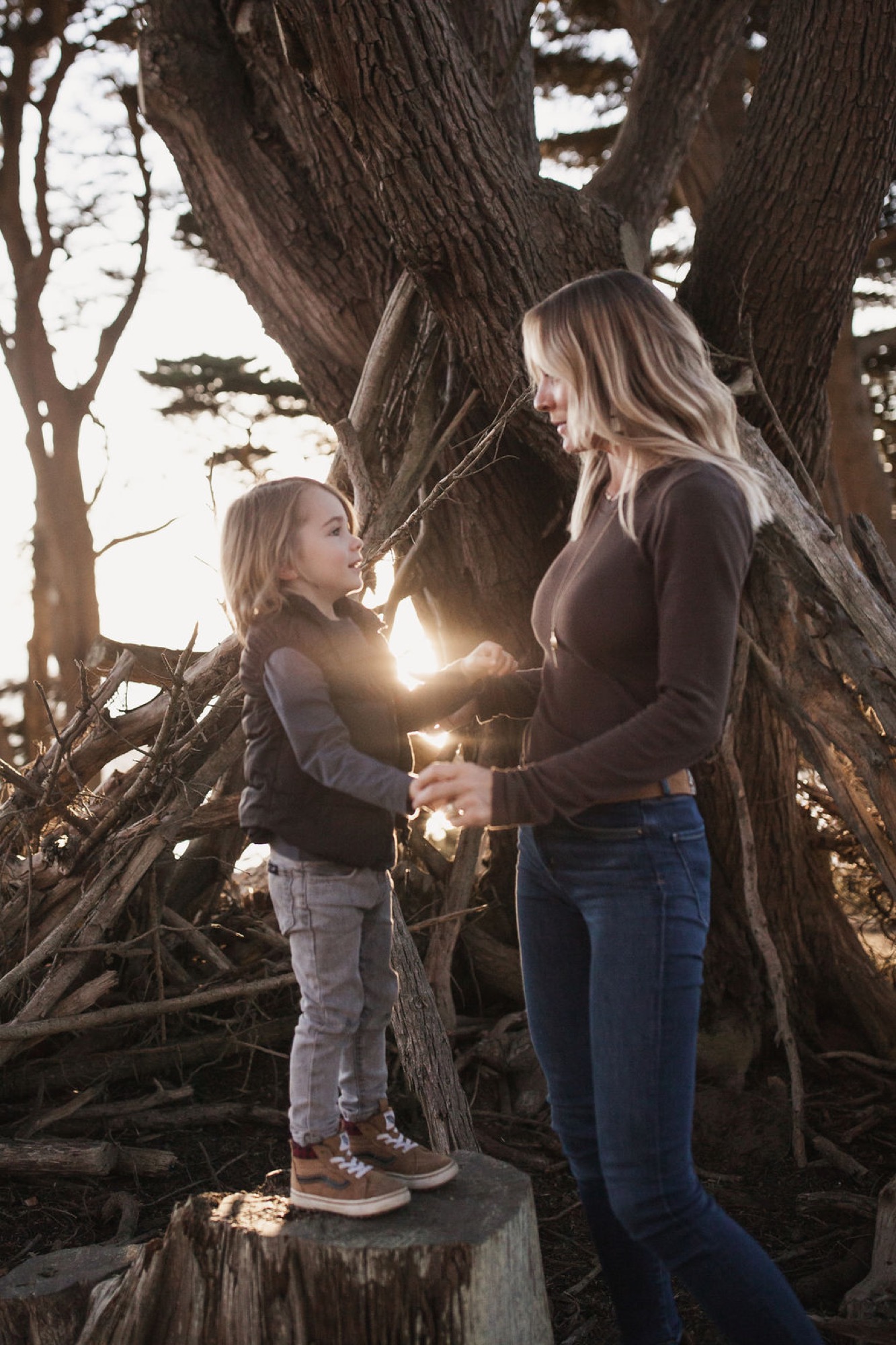 Mother and daughter stand face-to-face beneath a gnarled tree trunk, golden sunlight streaming between them as the younger child gazes upward with wonder