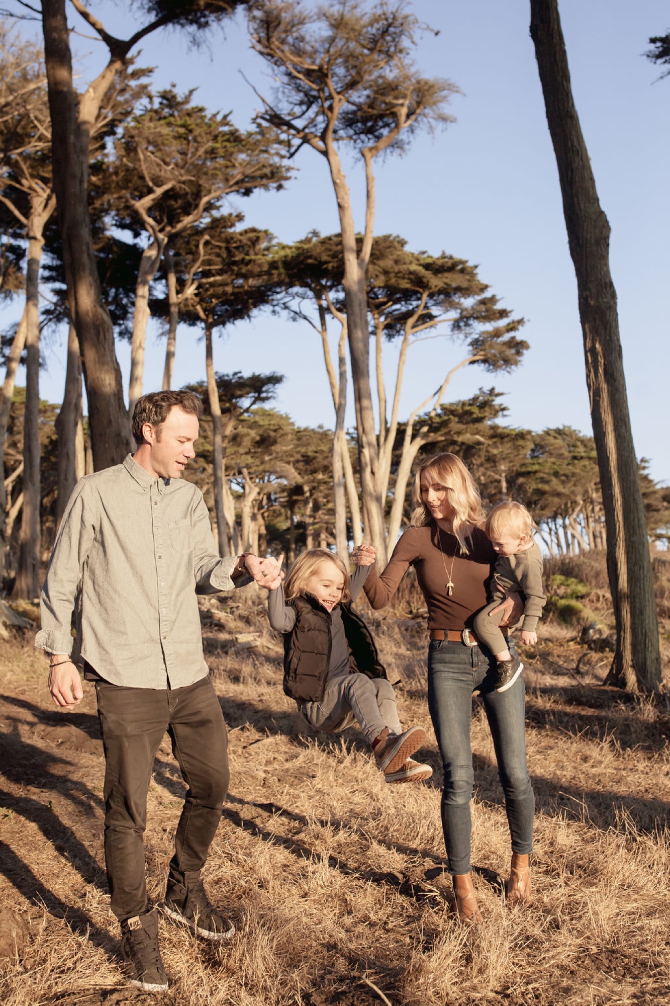 Family walks together through Lands End's iconic windswept cypress grove, dappled sunlight casting long shadows across the sandy trail