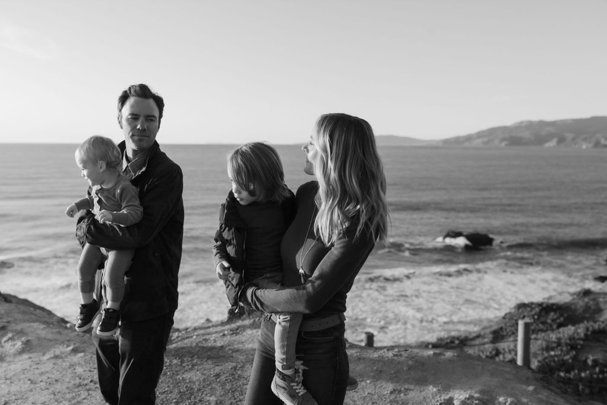Family of four stands together on a windswept coastal bluff, the father holding their youngest while the mother and older child lean in close, ocean and distant hills behind them