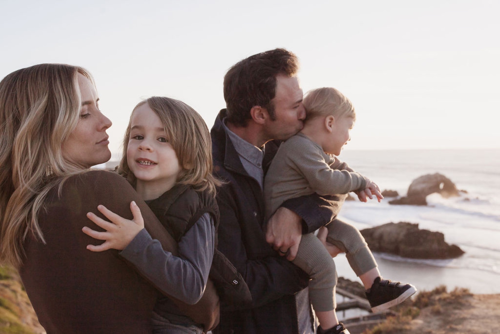 A family of four gazes toward the misty ocean horizon, the parents holding their young children close as soft coastal light bathes the rocky shoreline