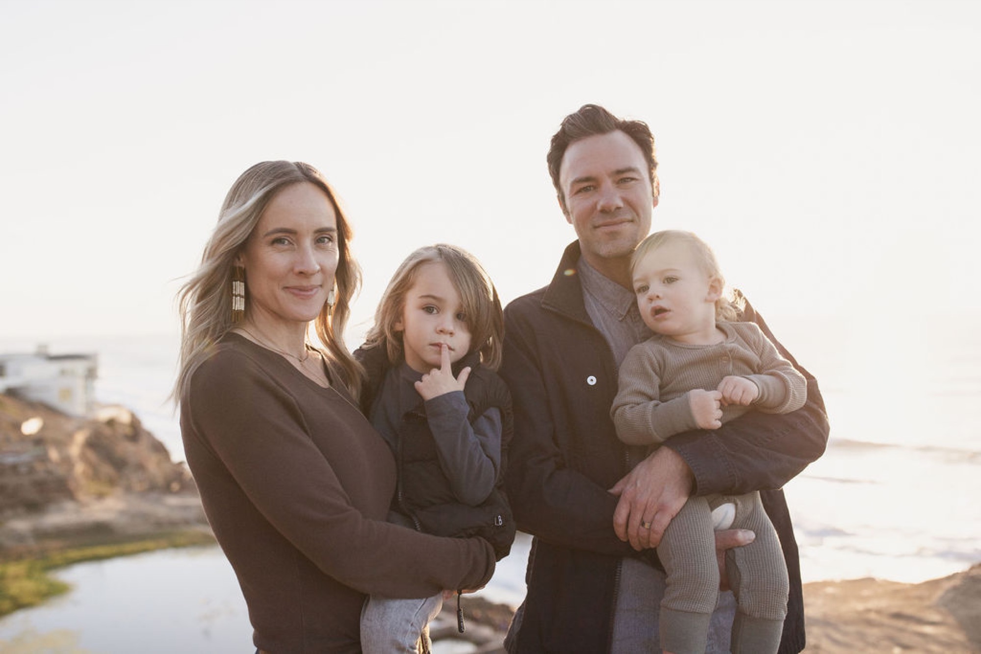 Family of four stands together at golden hour with calm water and rocky coastline behind them, all gazing toward the camera with gentle smiles