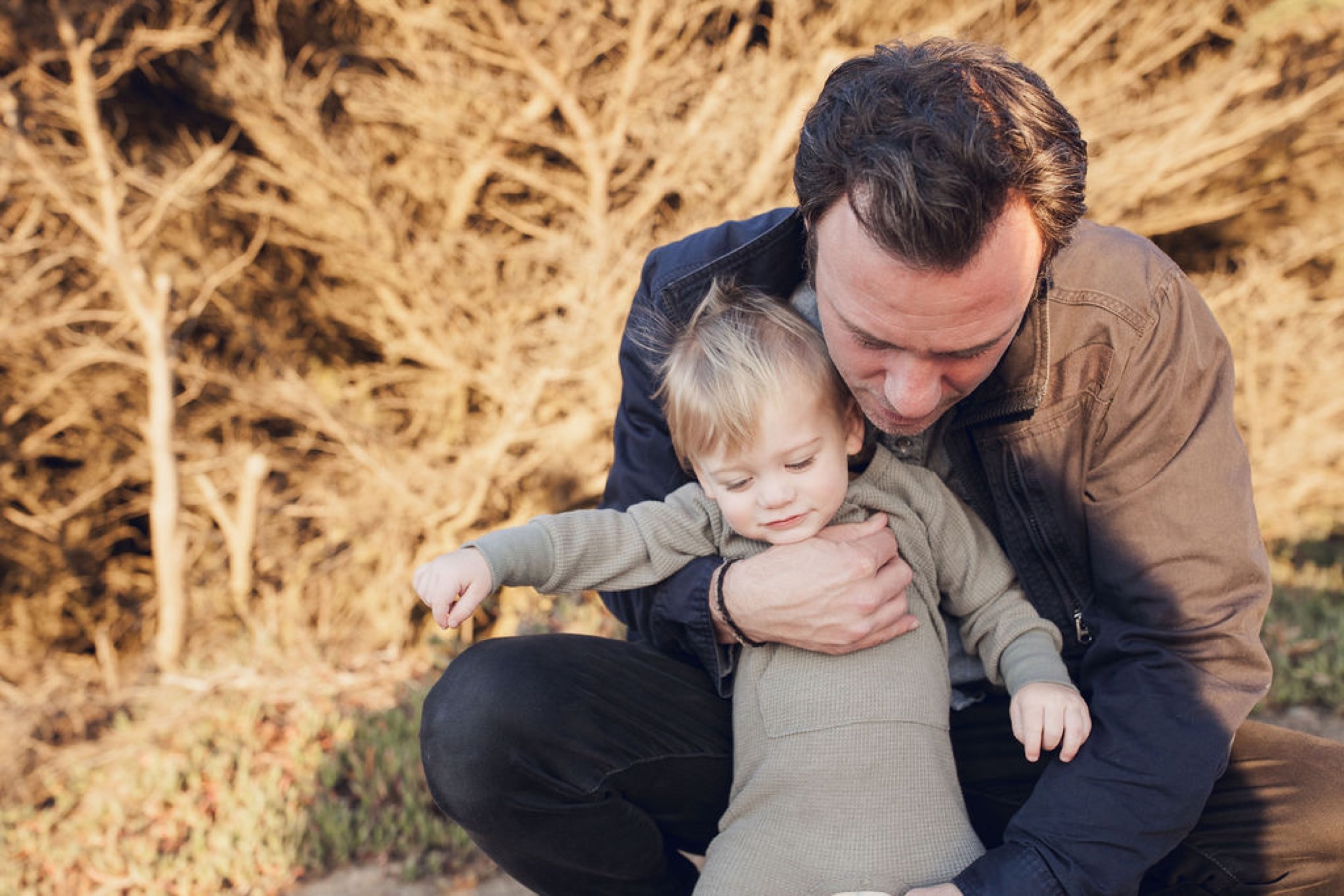 A father crouches beside his toddler son among dried grasses, gently supporting the child's outstretched arm as golden afternoon light filters across their faces