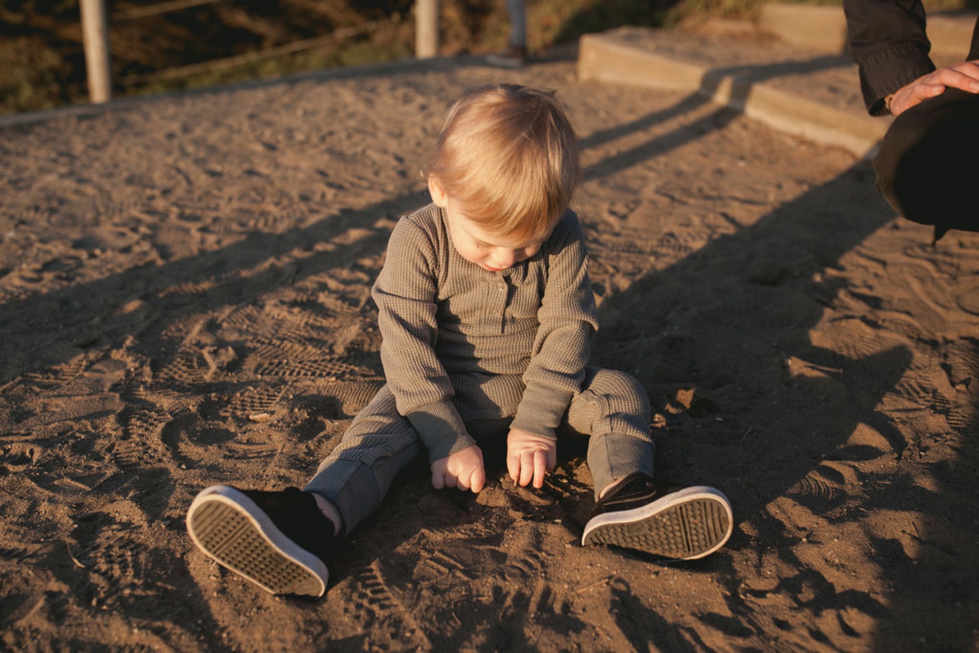 Toddler sits cross-legged on sandy ground in golden afternoon light, studying their sneakered feet with focused attention