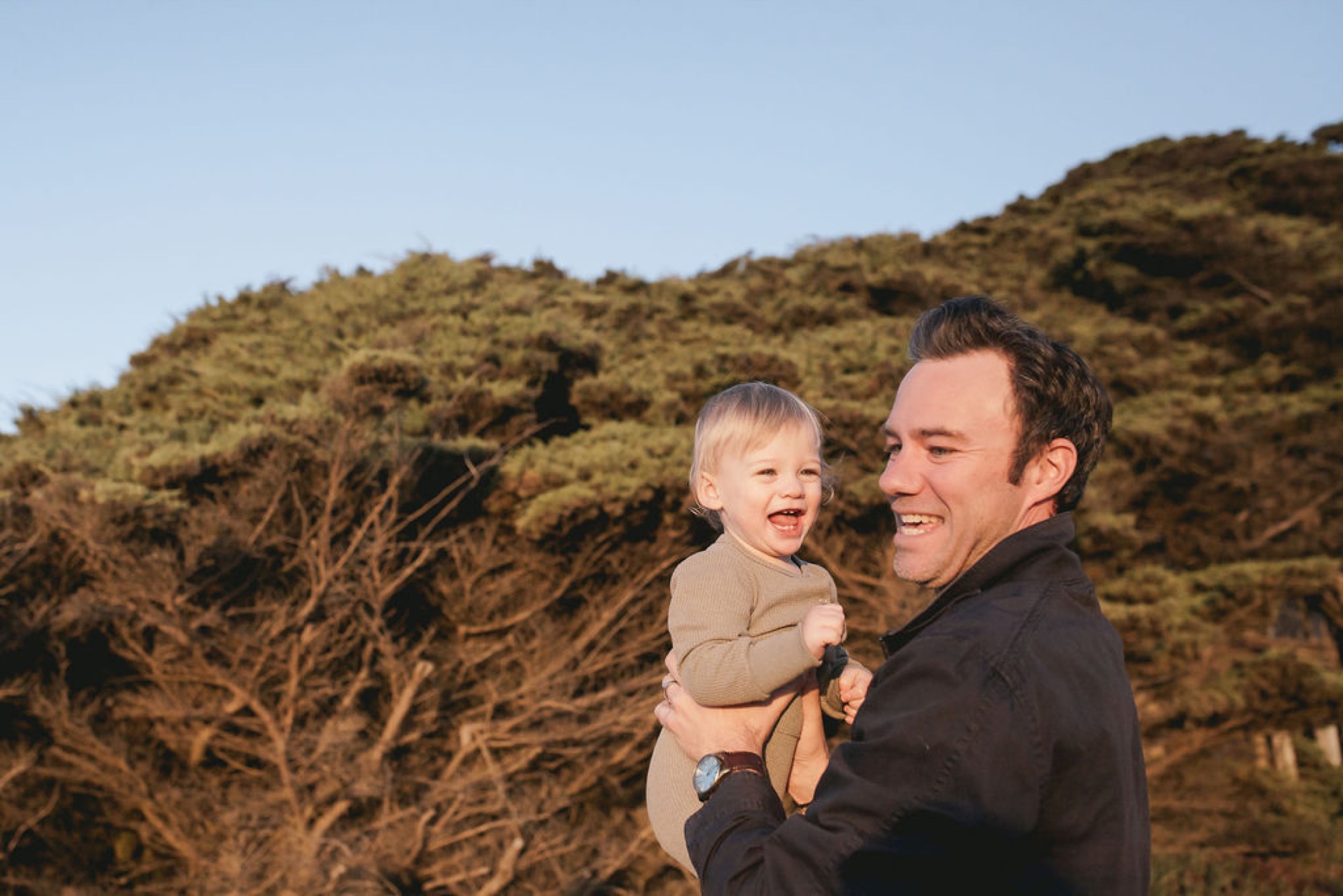 A man holds his laughing toddler against his chest while standing on a hillside with golden brush and bare branches behind them