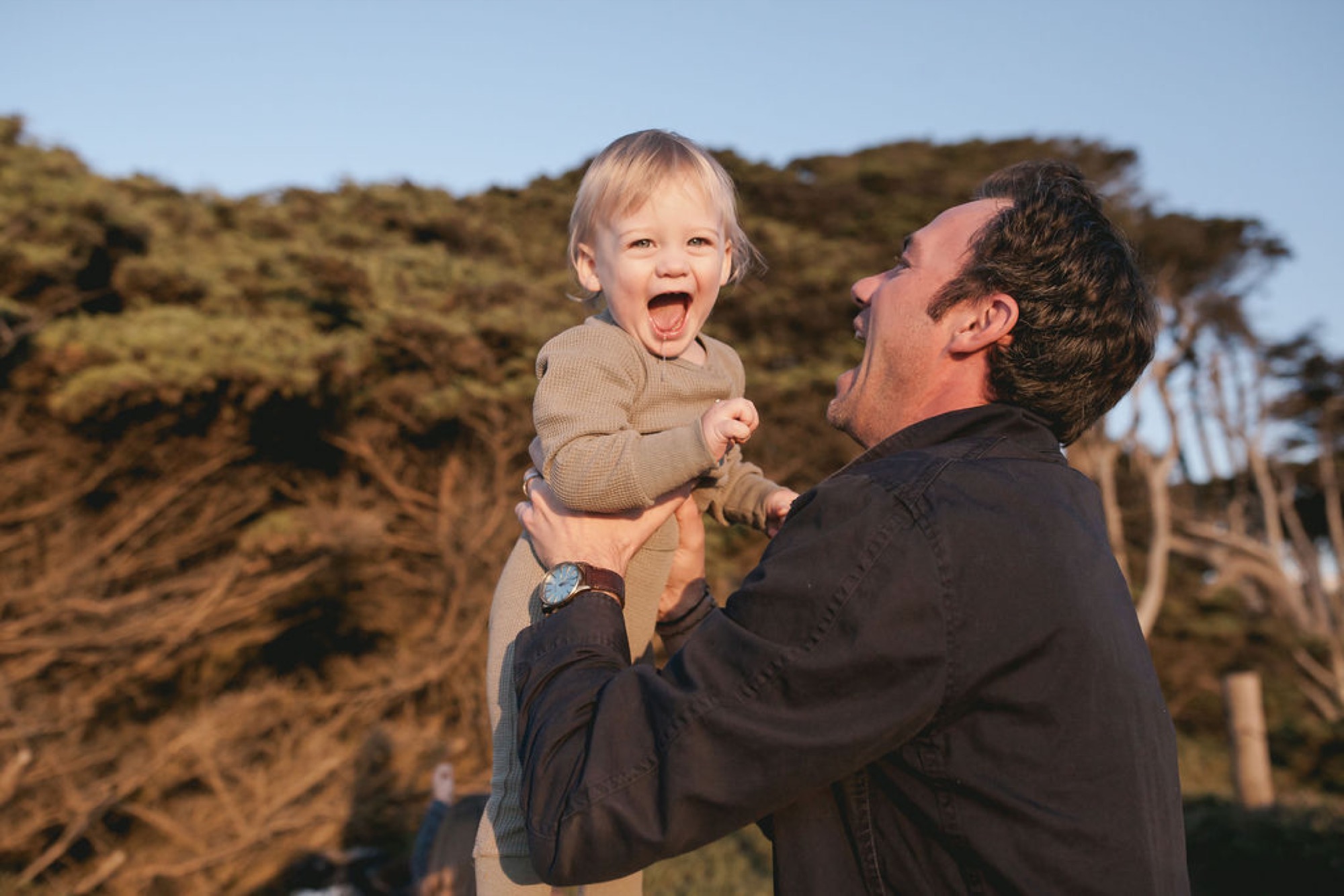 A man lifts a blonde toddler high against his chest, both laughing with open mouths as golden hillside vegetation glows behind them