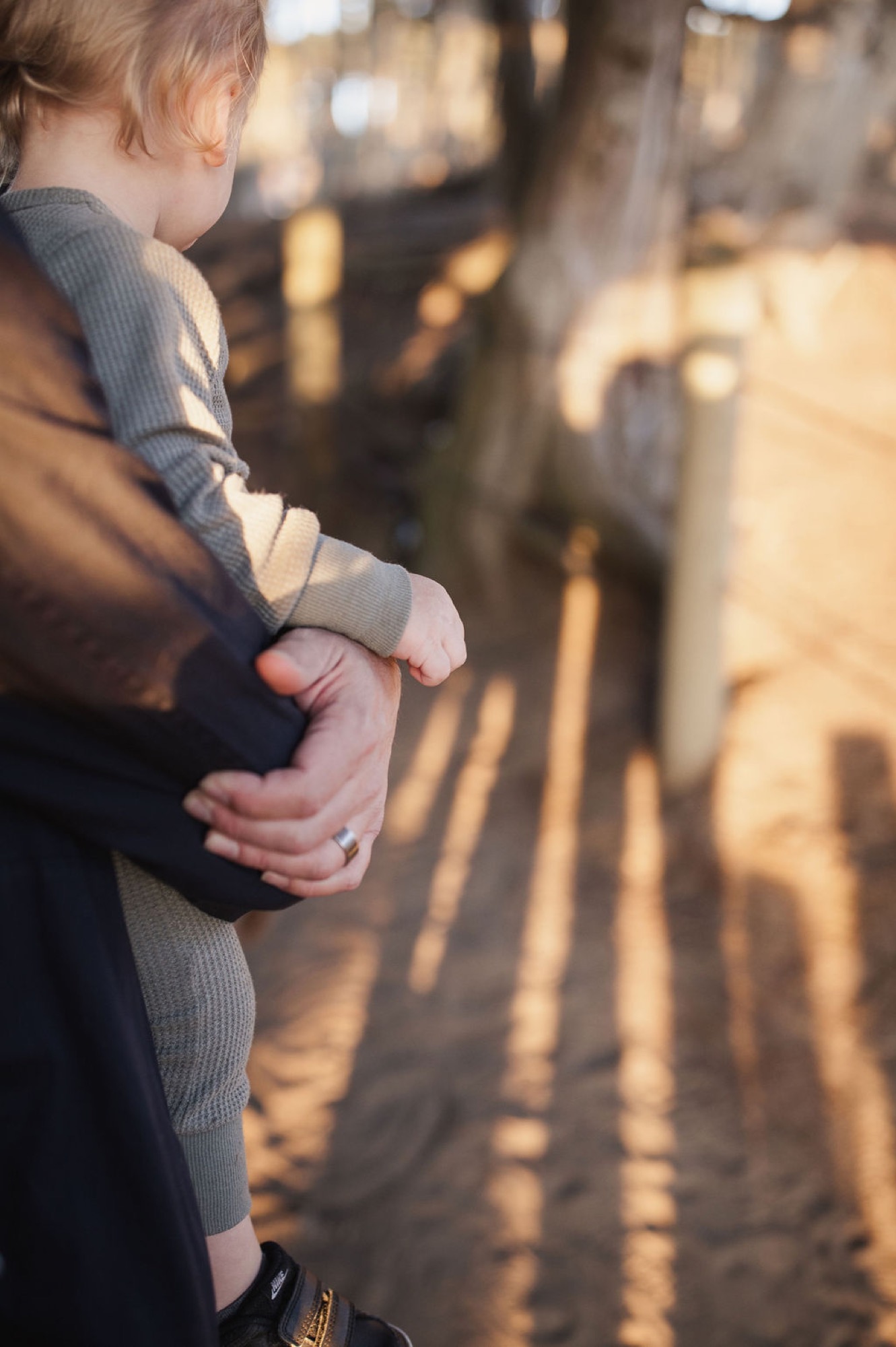 A child in gray clothing stands at the edge of a sun-dappled coastal path, their small hand held gently by an adult, golden afternoon light casting long shadows across the ground