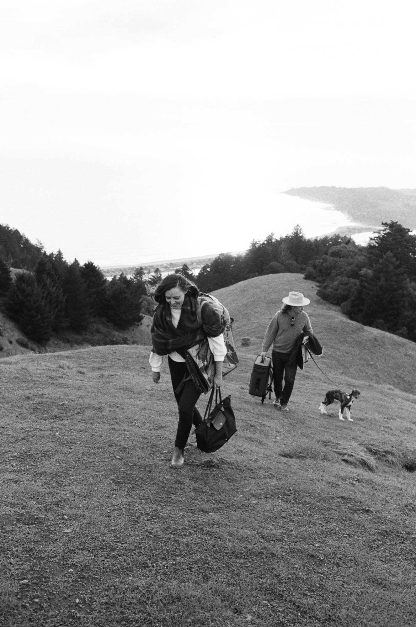 A woman in a dark jacket strides forward down a gravel path, smiling broadly, while two companions and a small dog follow across rolling hillsides with a distant bay visible