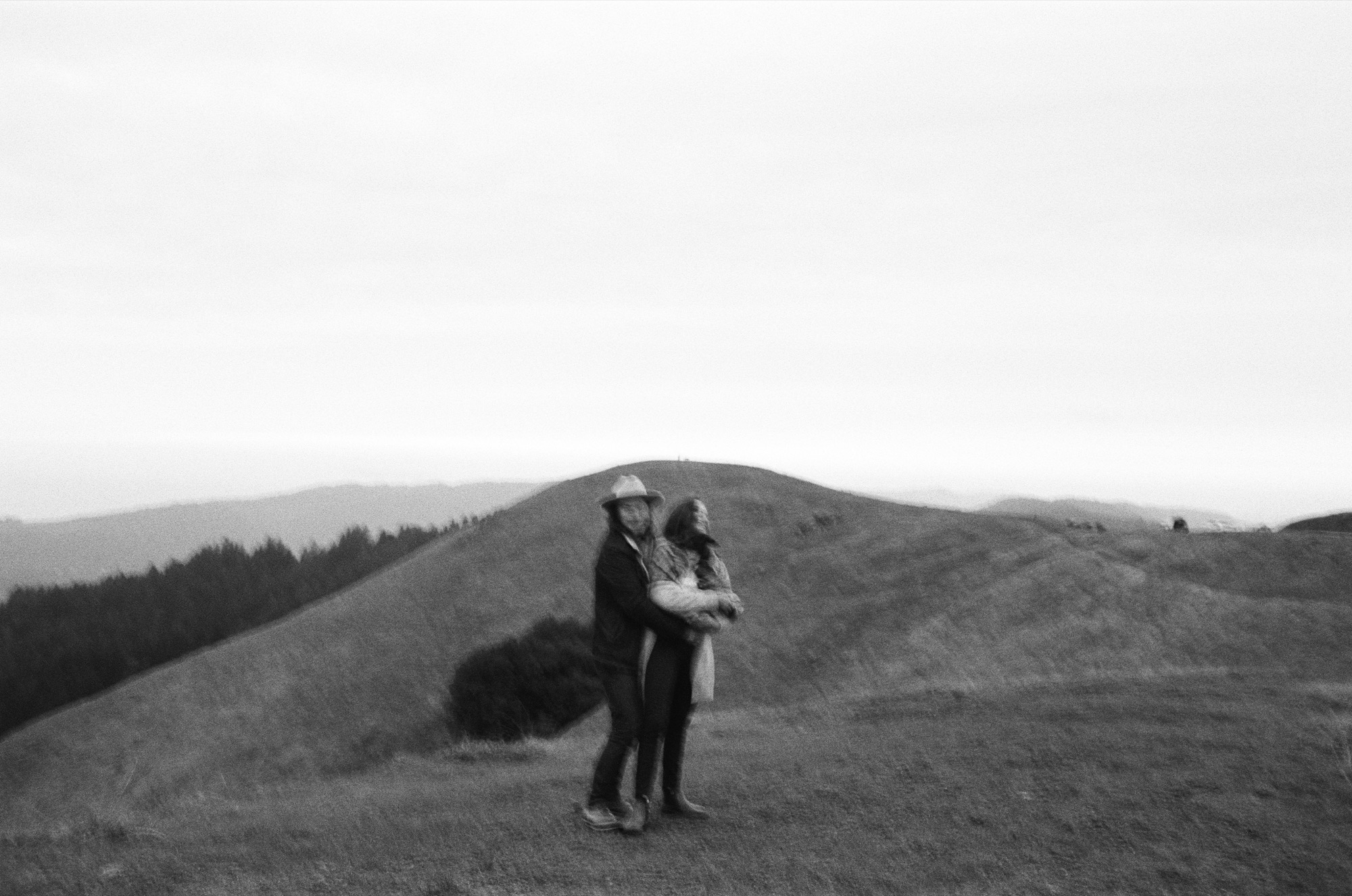A couple stands embraced on a windswept hilltop, their silhouettes framed against distant forested ridges under overcast skies