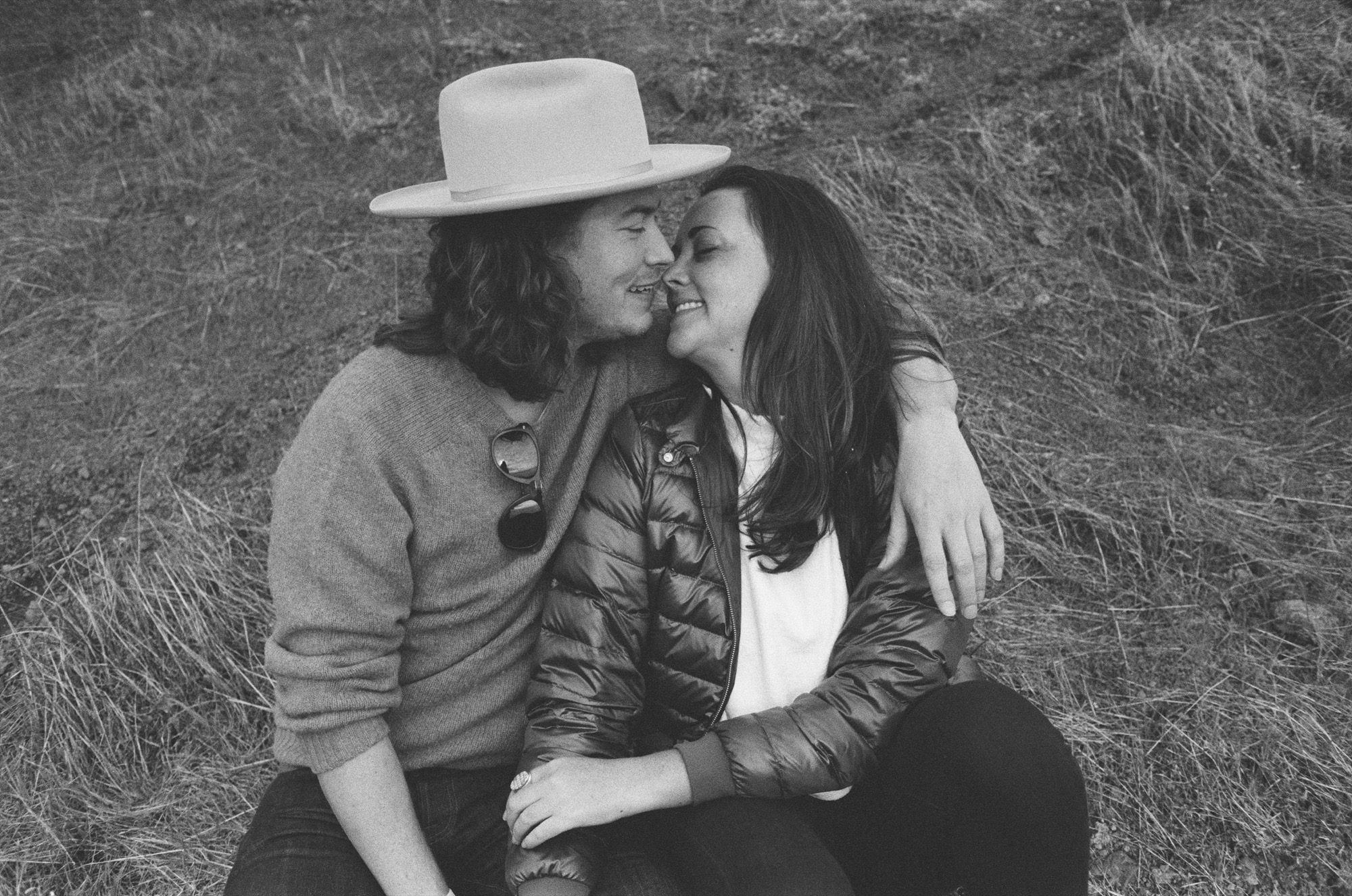 Two people sit close together on dry grass, sharing an intimate moment with genuine smiles and arm-in-arm embrace