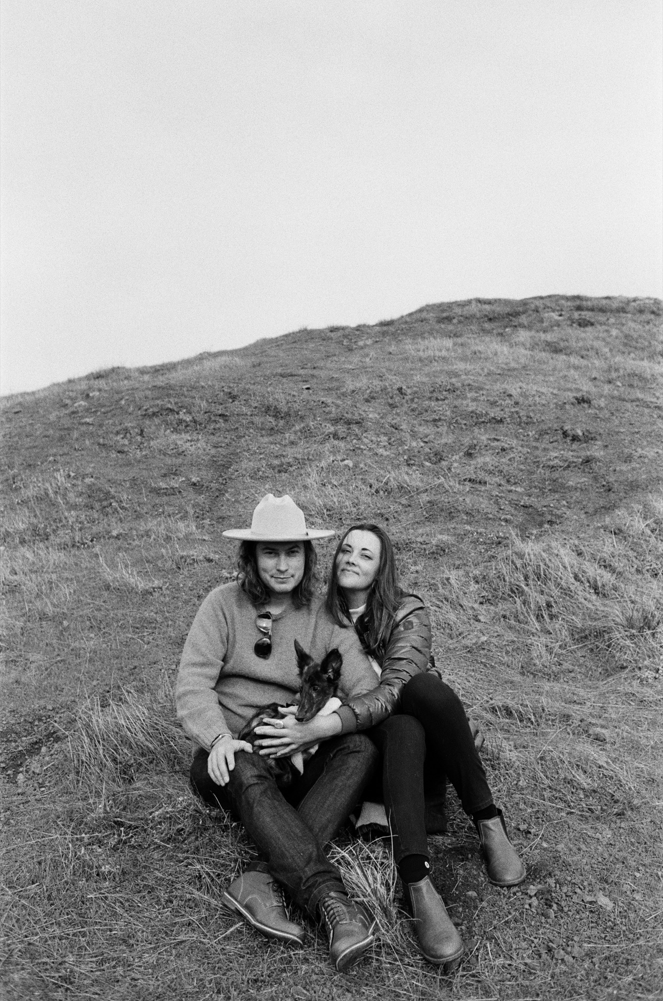 Two people sit close together on a grassy hillside, one wearing a cowboy hat, both gazing upward while holding a small dark dog