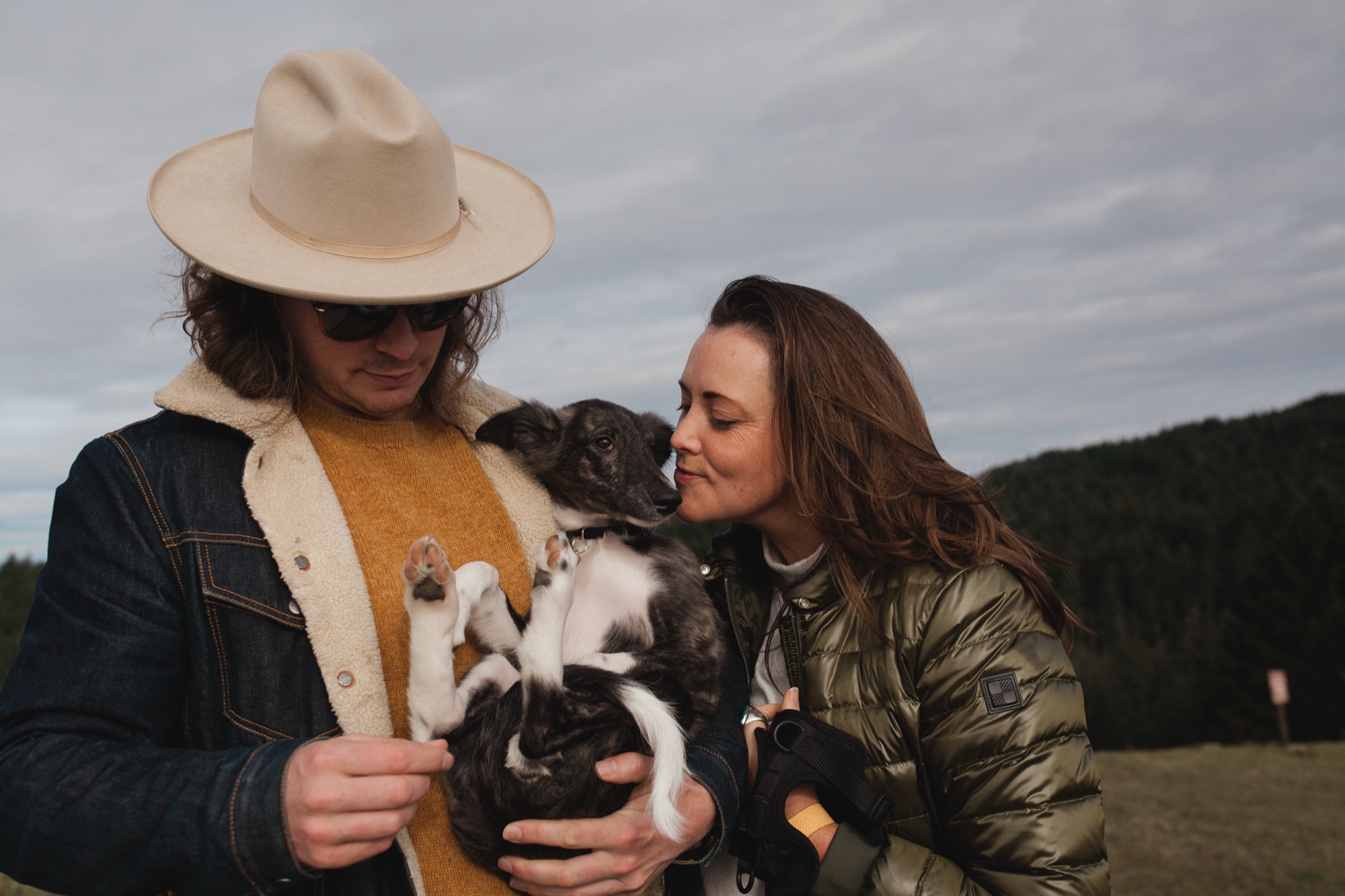 A man in a cream cowboy hat and sherpa jacket holds a black and white puppy while a woman in an olive puffer jacket gazes at the dog in an open meadow