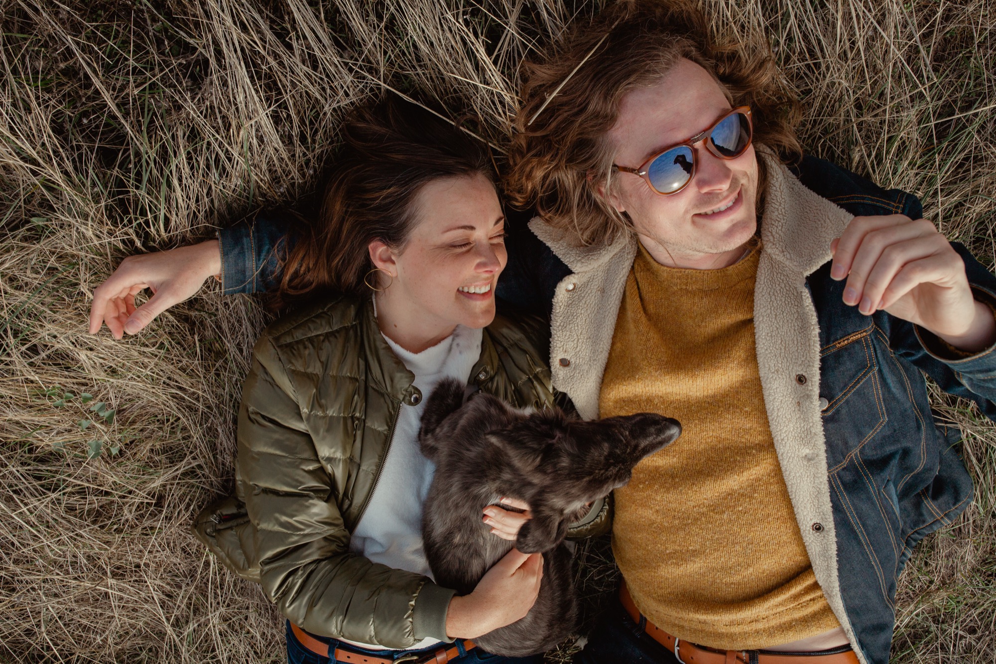 Two people lie on dry grass, smiling at each other while holding a black cat between them in bright sunlight