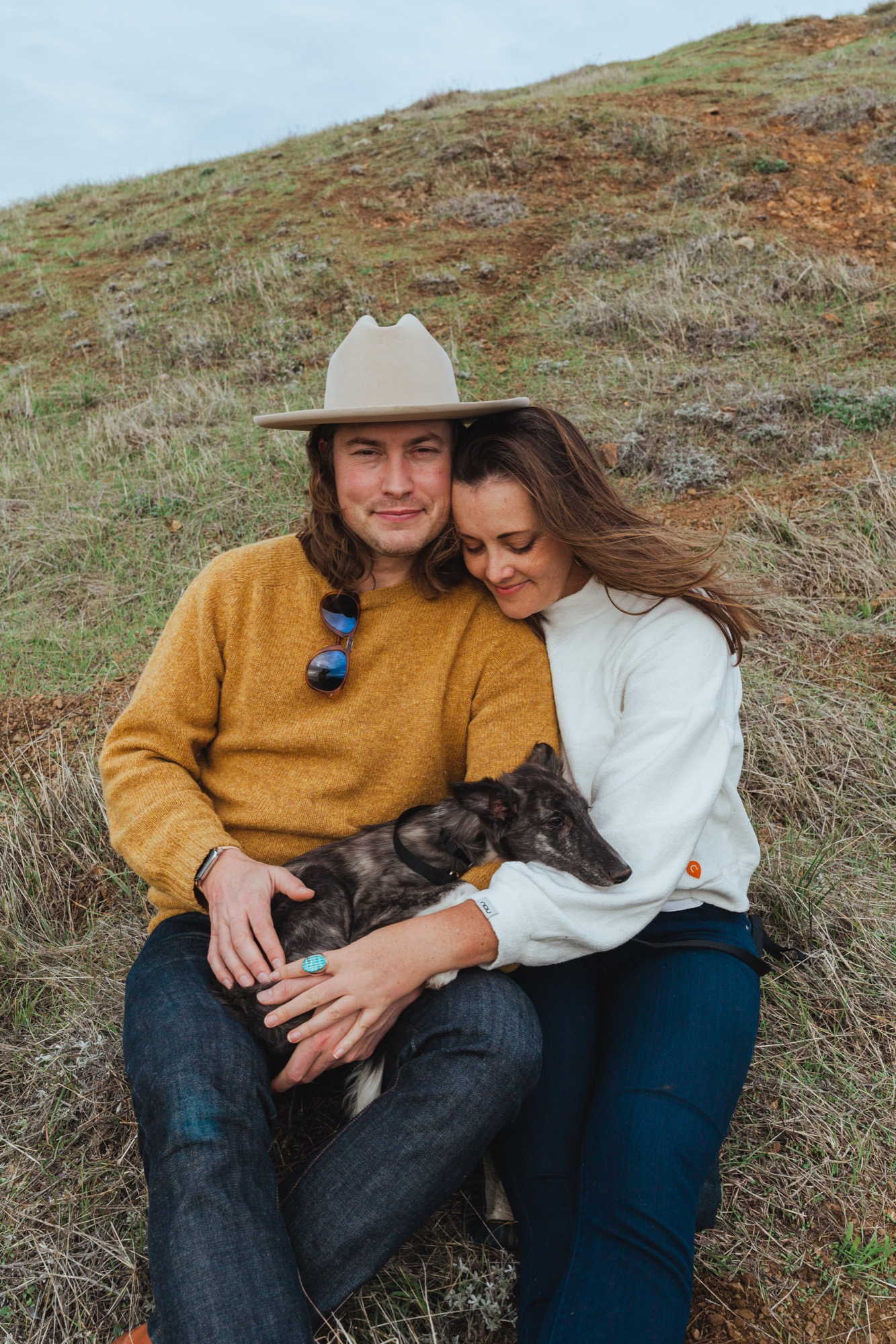 A couple sits close together on a grassy hillside, the man wearing a cream cowboy hat and mustard sweater, both smiling softly while holding their dark dog