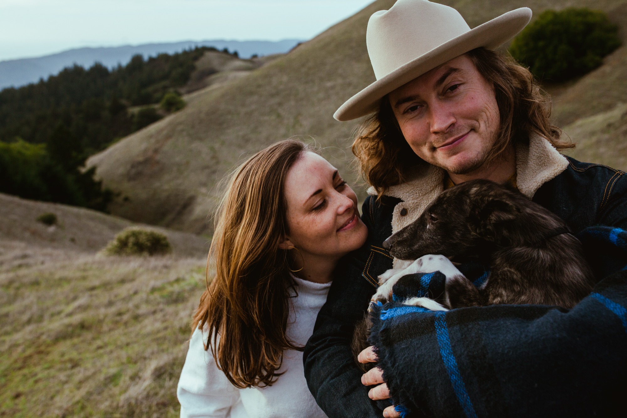 A couple smiles warmly while holding their dark puppy wrapped in a blue plaid blanket against rolling golden hillsides