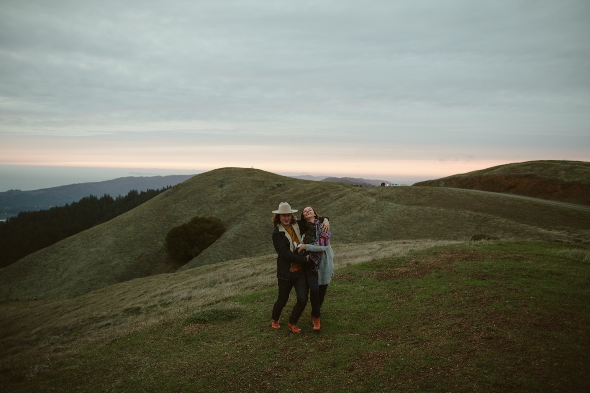 Two women embrace on a rolling hillside at dusk, looking skyward together with genuine joy and connection