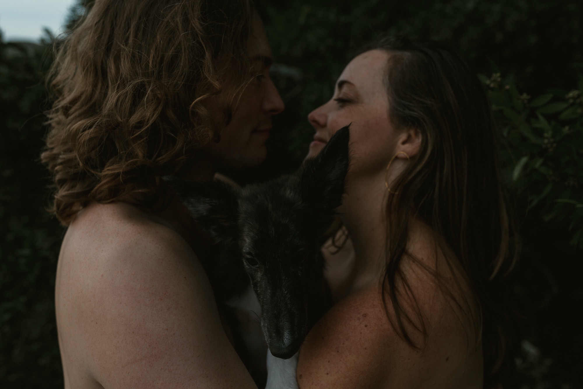 Two women embrace intimately outdoors surrounded by dark green foliage, their faces close in a tender moment