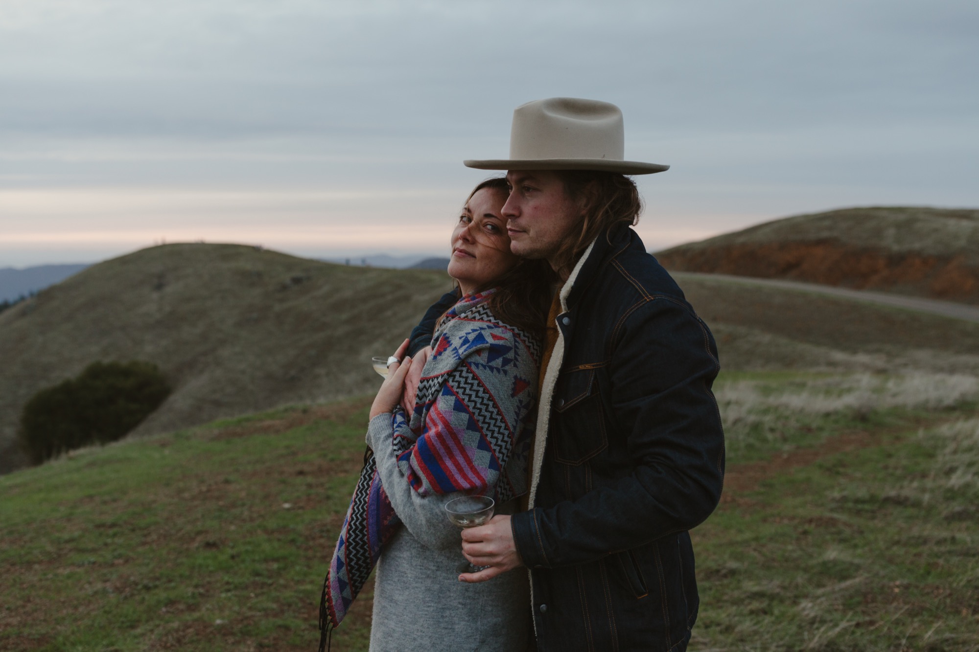 A couple stands embraced on a windswept hillside, gazing toward distant mountains beneath overcast skies, wrapped in patterned textiles and denim