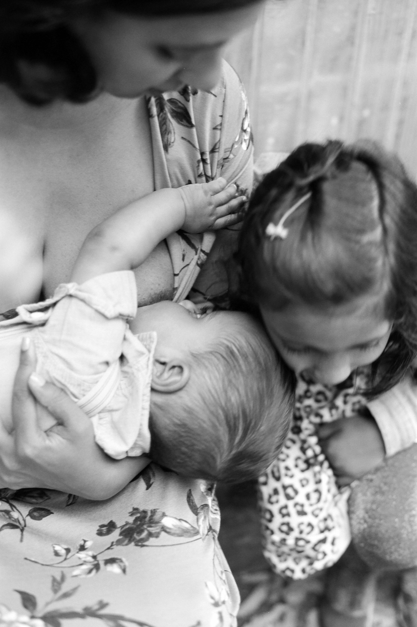 Two young children in patterned clothing sit closely together on floral fabric, their heads bent in gentle connection and focused attention