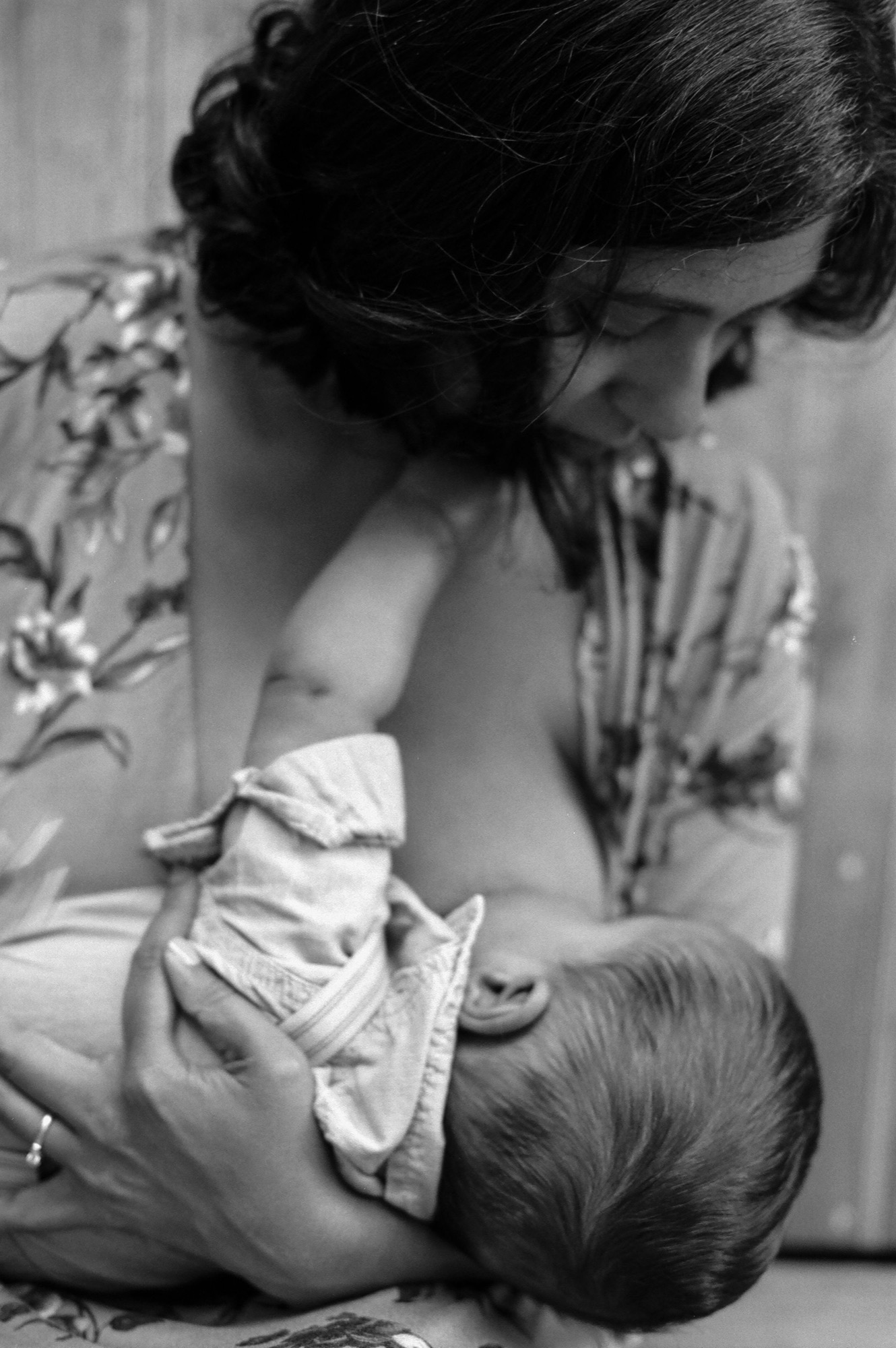 A mother tenderly holds her infant wrapped in white cloth, gazing downward with gentle affection in soft natural light