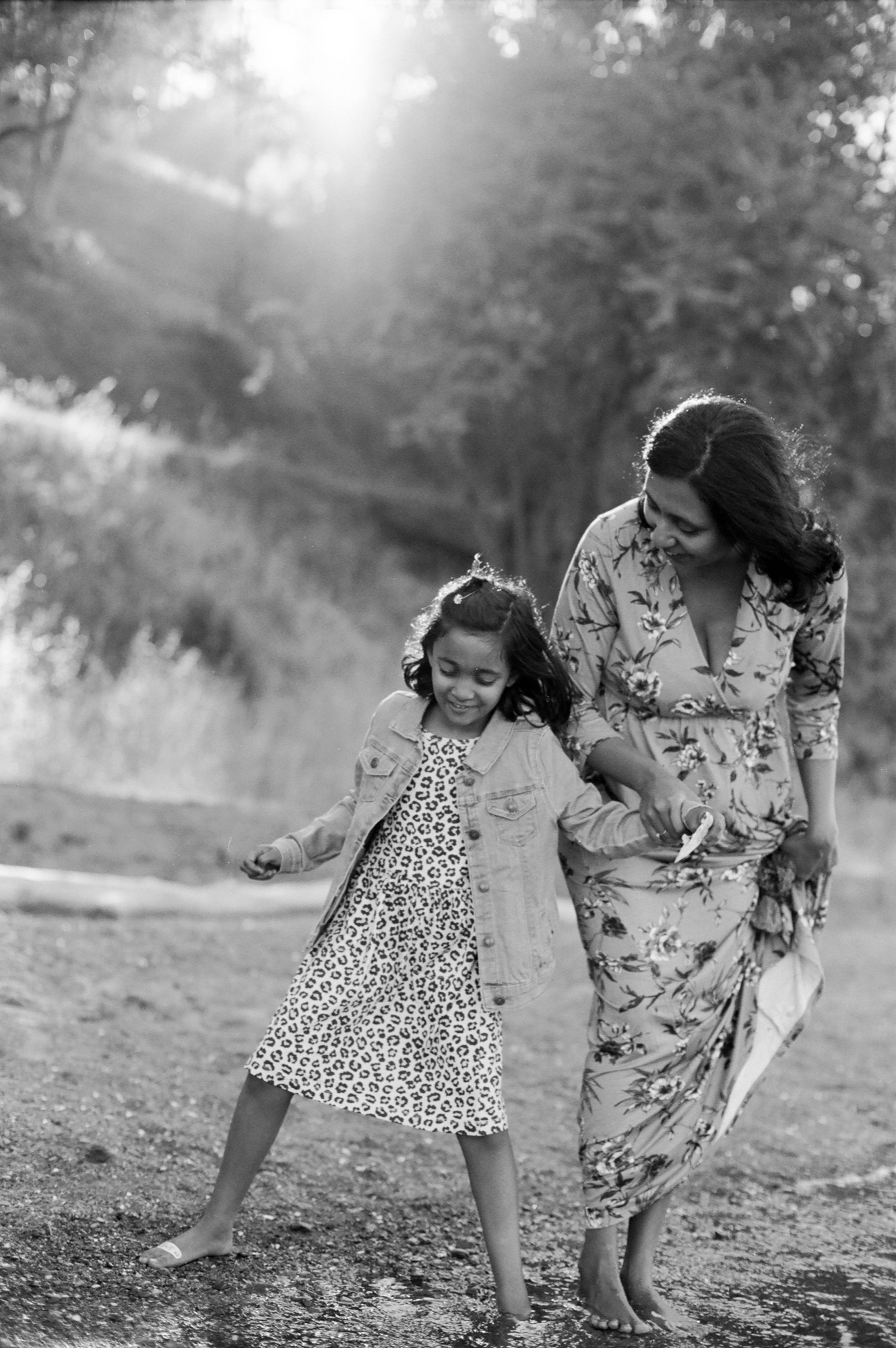 A mother and young daughter walk barefoot on a mountain road, the girl's arm extended joyfully as sunlight filters through towering forest