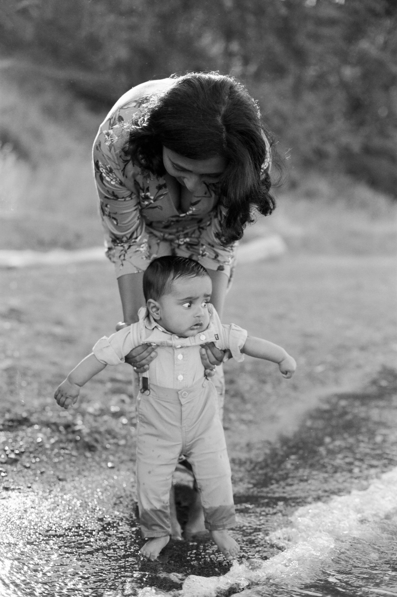 A woman guides her toddler through shallow water, both absorbed in the moment as backlit waves lap at their feet
