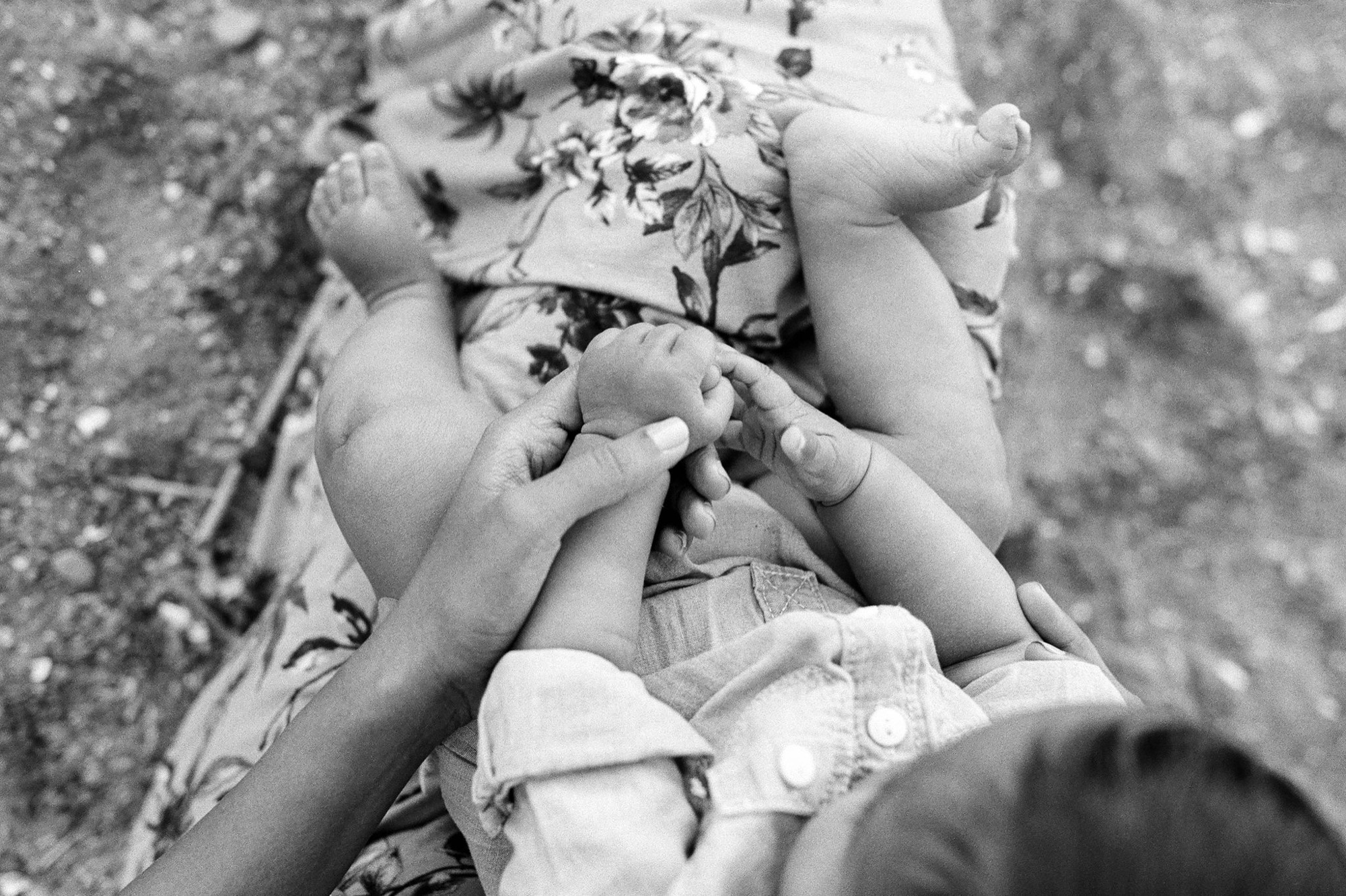 A parent's hands gently guide a young child's fingers as they sit together on sandy ground, captured in soft black and white