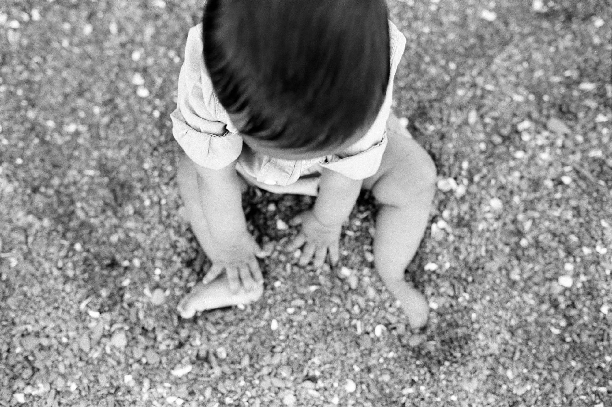 Barefoot toddler in white shirt crouches on sandy beach, dark hair obscuring face as small hands explore the grainy shore
