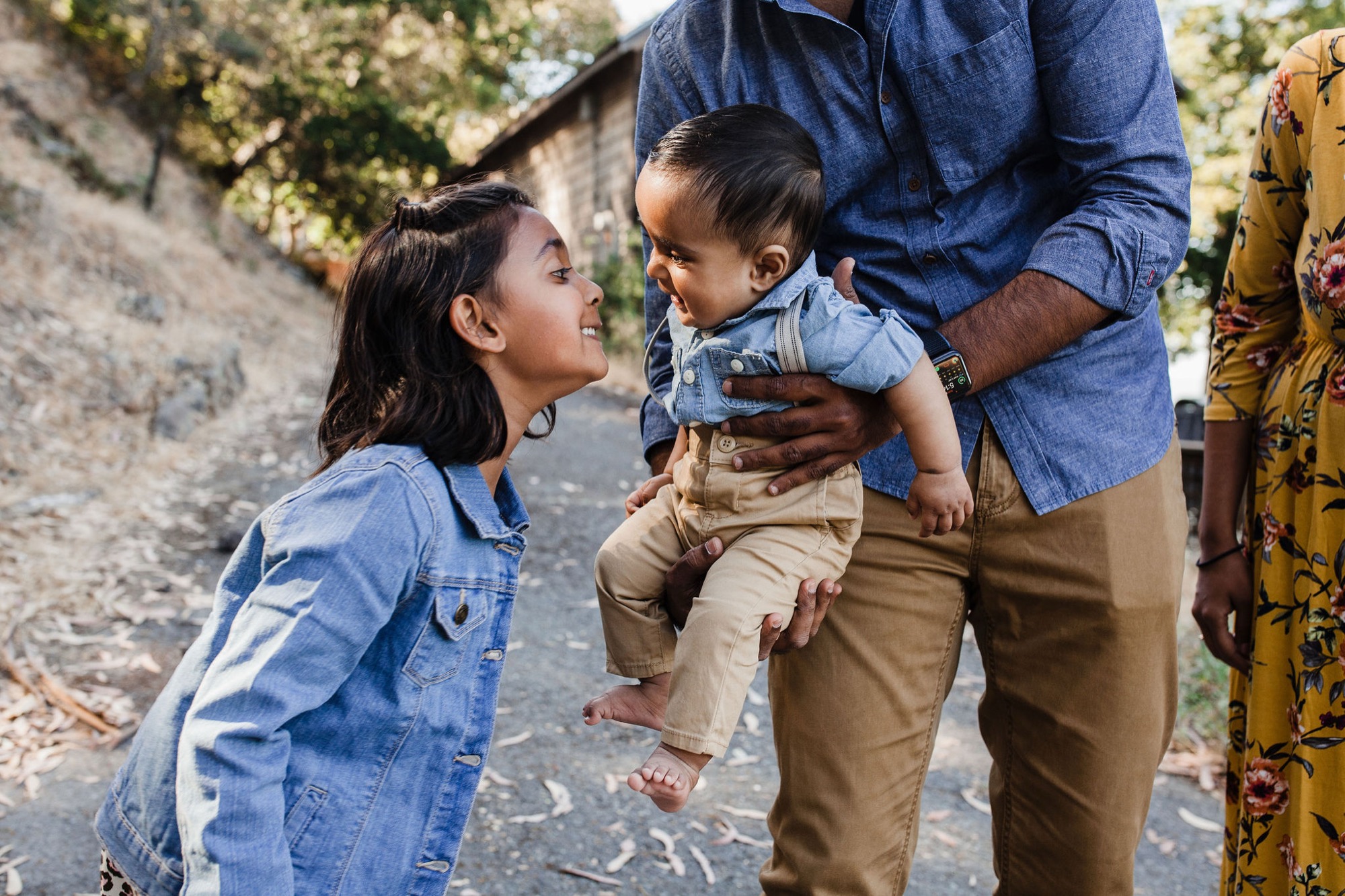 An older sister beams at her toddler brother while their father holds him on a sunlit cobblestone path lined with weathered stone walls