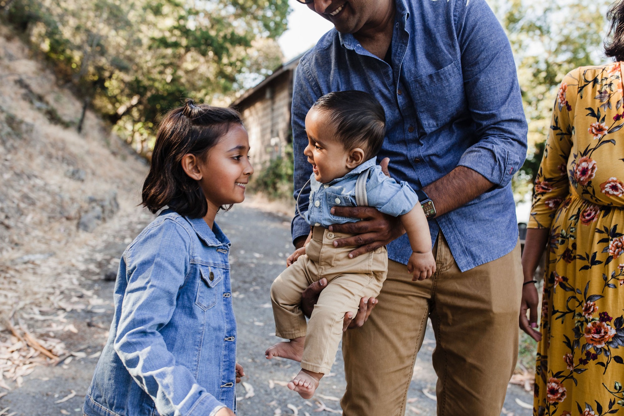 A father holds his toddler son while his older daughter smiles up at them on a sunlit stone pathway, creating a warm moment of family connection