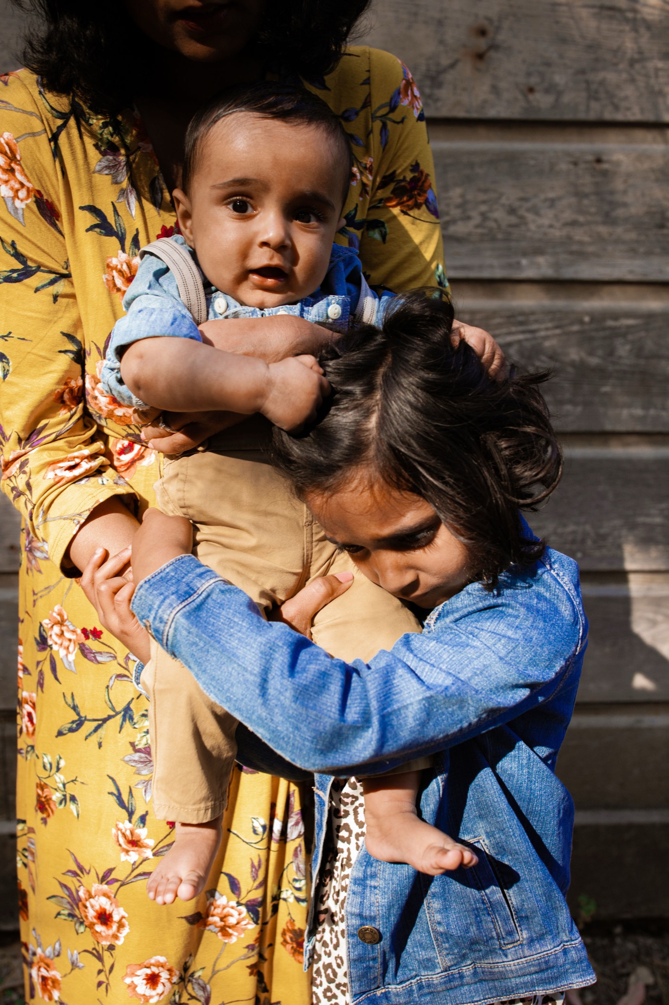 A toddler in a blue checkered shirt gazes curiously while an older sister hugs from behind, both held by their mother in a yellow floral dress against weathered wooden steps