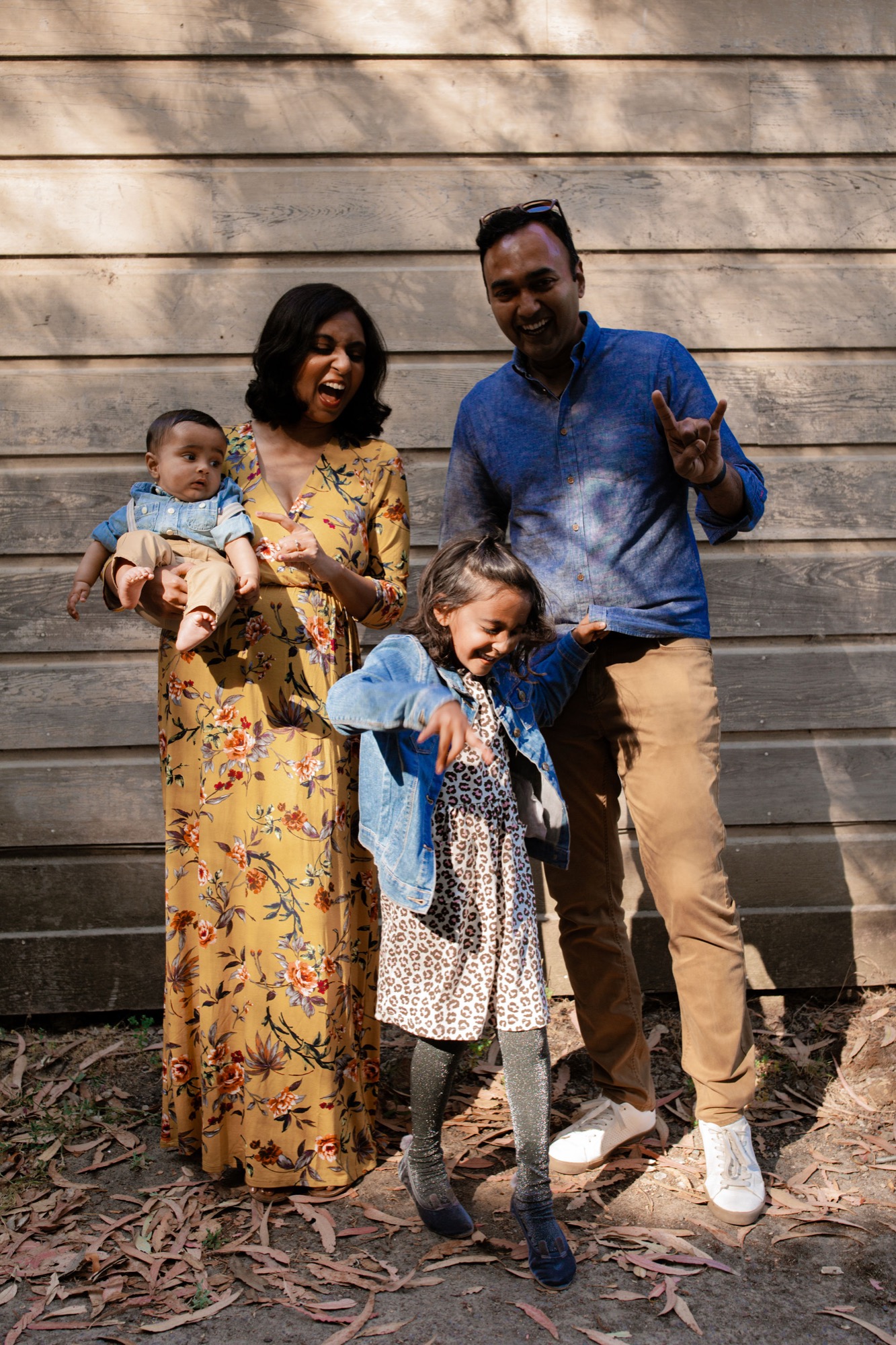 A joyful family of four laughs together against weathered wooden siding, with dappled sunlight casting shadows across their faces and the ground