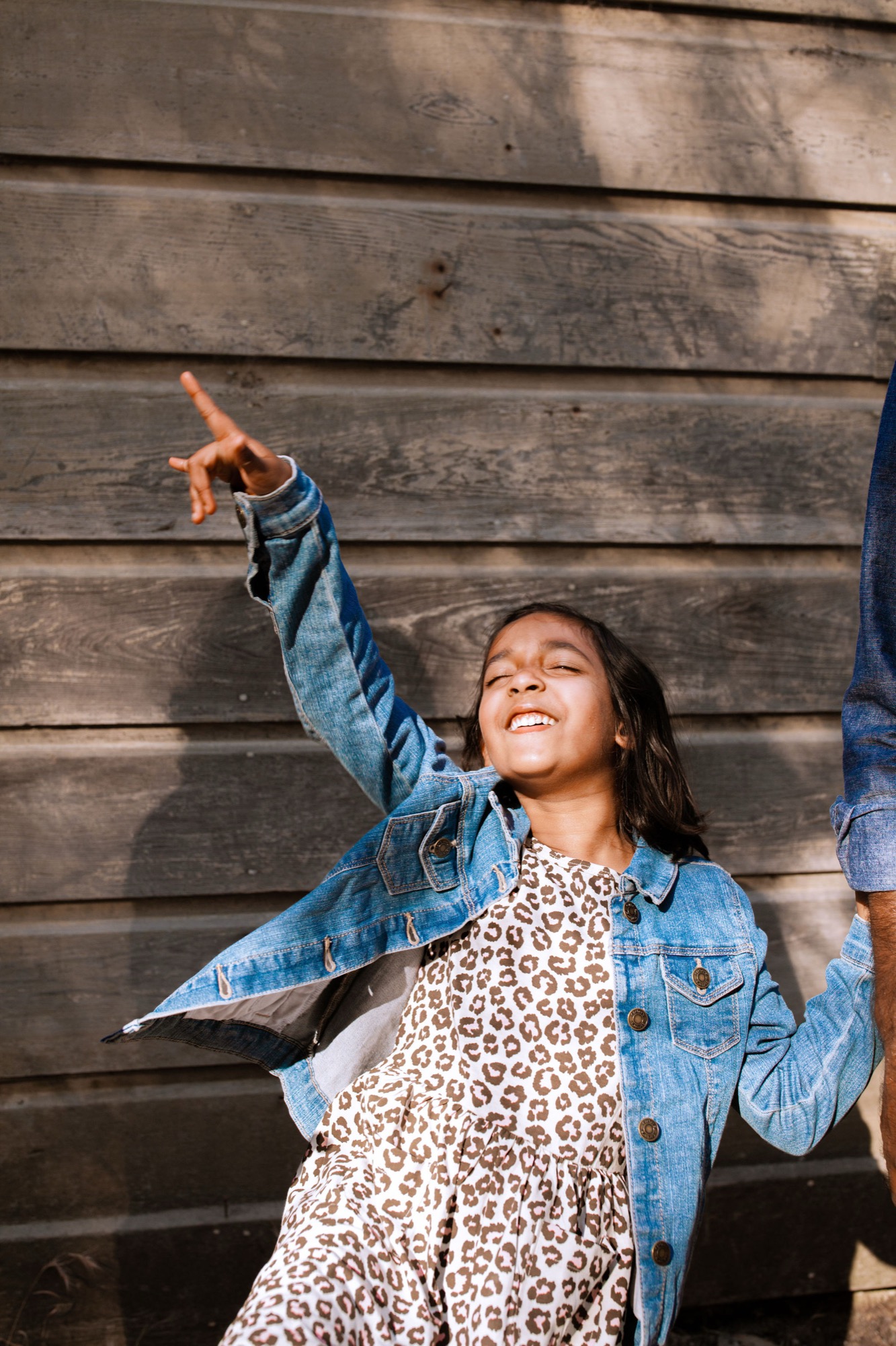 A young girl in a denim jacket and leopard-print dress throws her arm up joyfully against dark wooden shiplap, beaming with uninhibited happiness