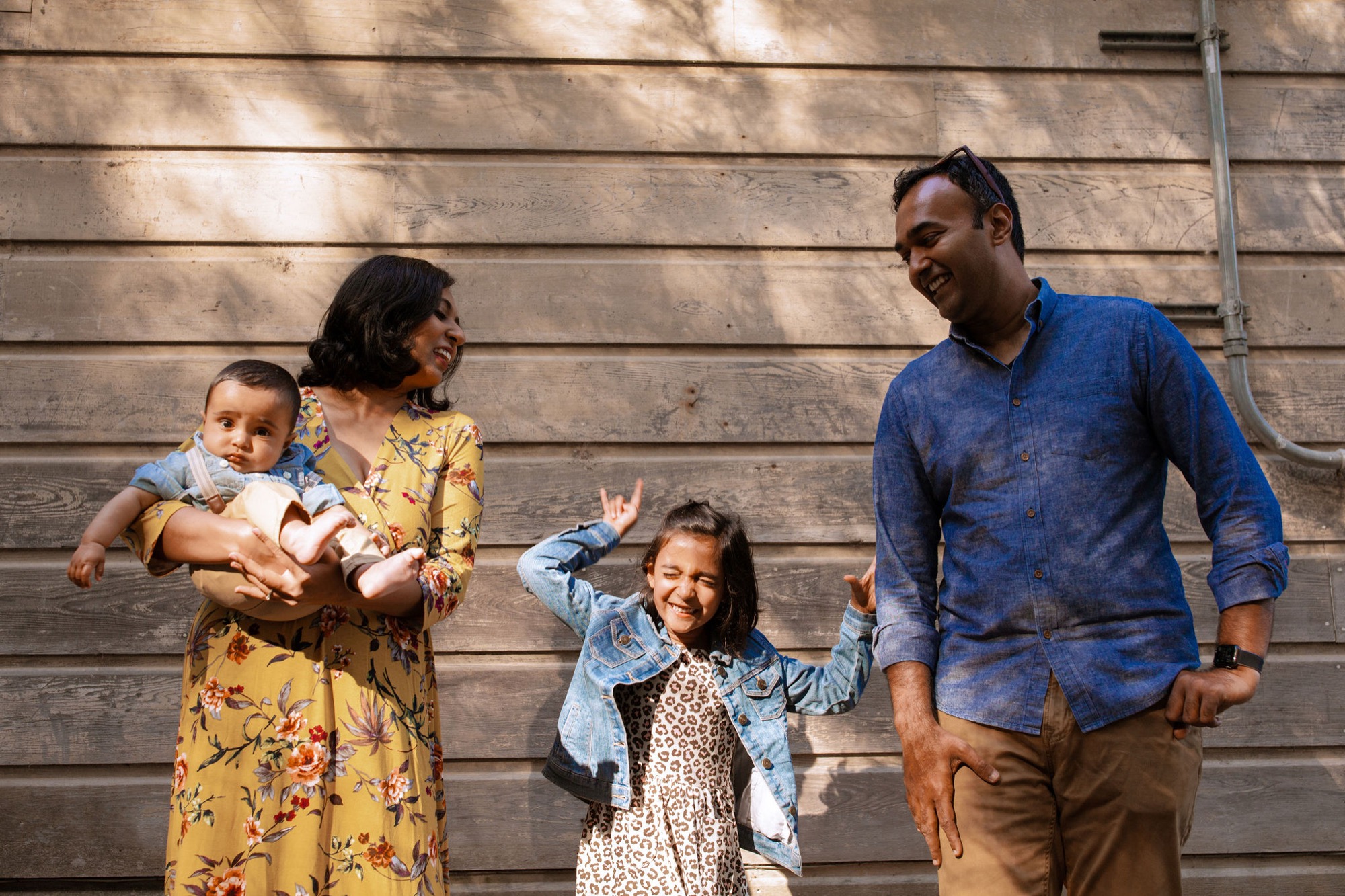 A joyful family of four stands against weathered wooden siding, the father and daughter beaming while the mother holds an infant in soft afternoon sunlight