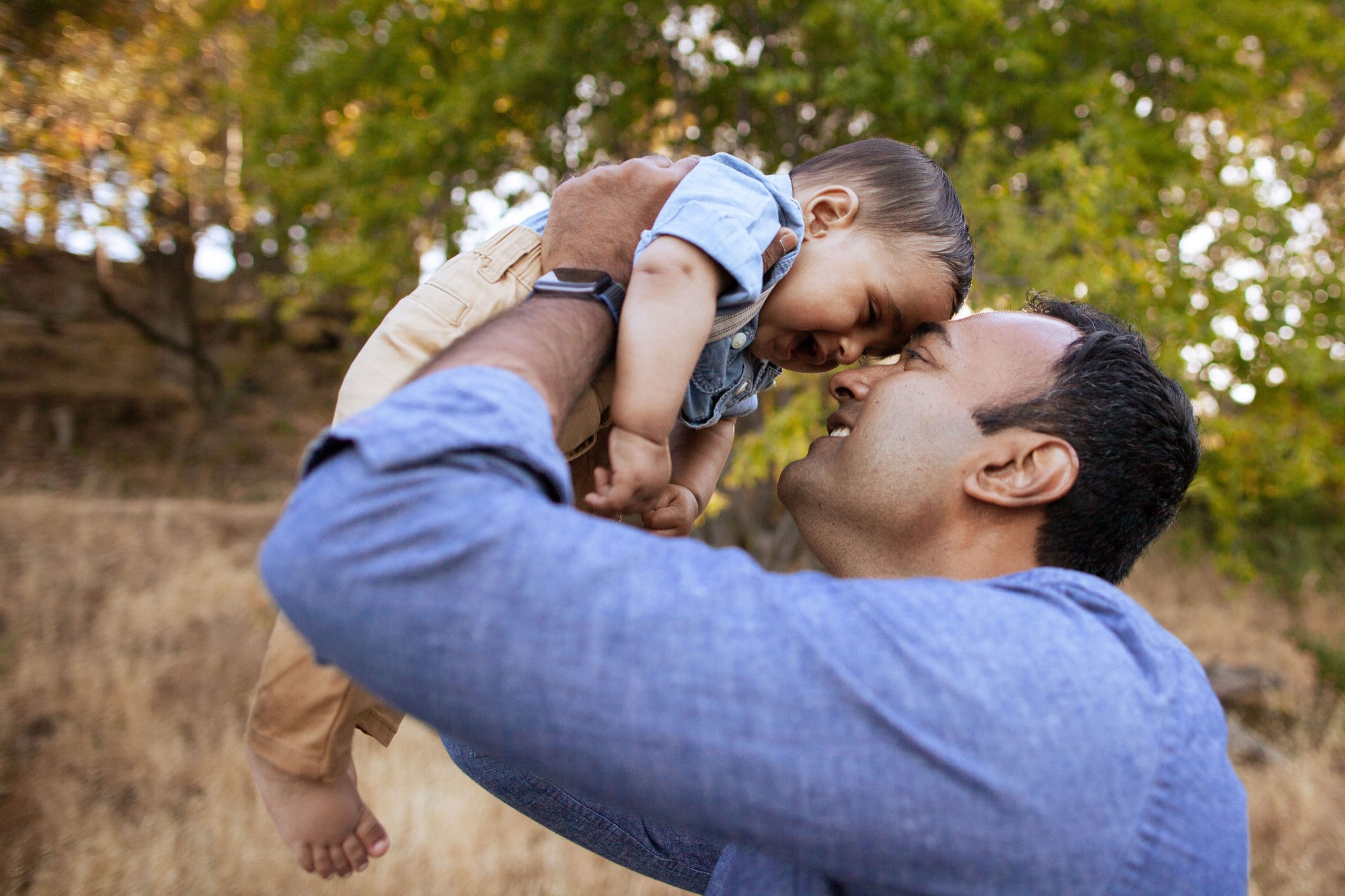A father lifts his young son high in the air, both smiling with joy as dappled sunlight filters through the green canopy above them