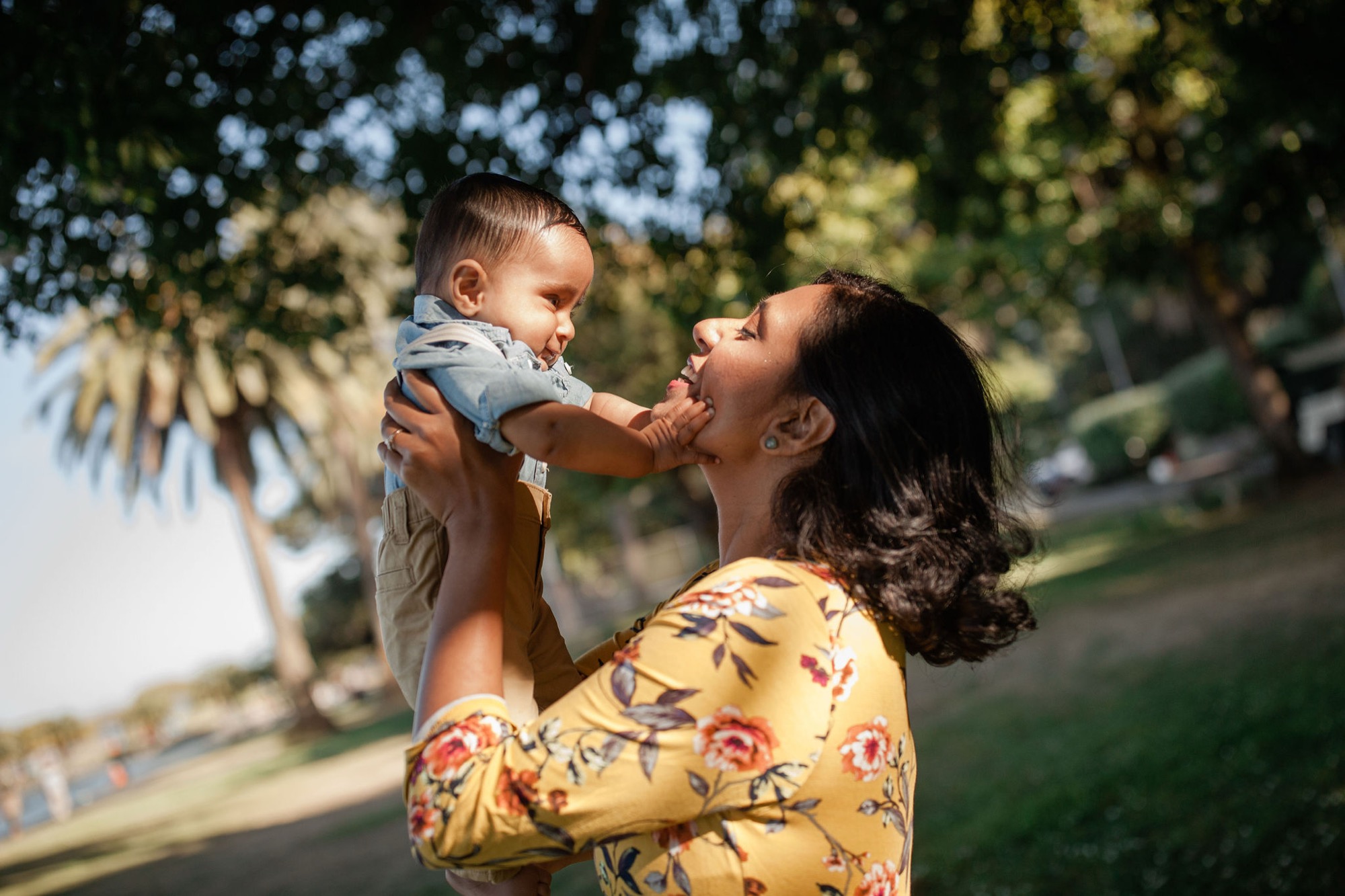 A mother in a yellow floral dress lifts her smiling toddler toward the sky in dappled sunlight, their joy radiating as palm fronds frame the moment