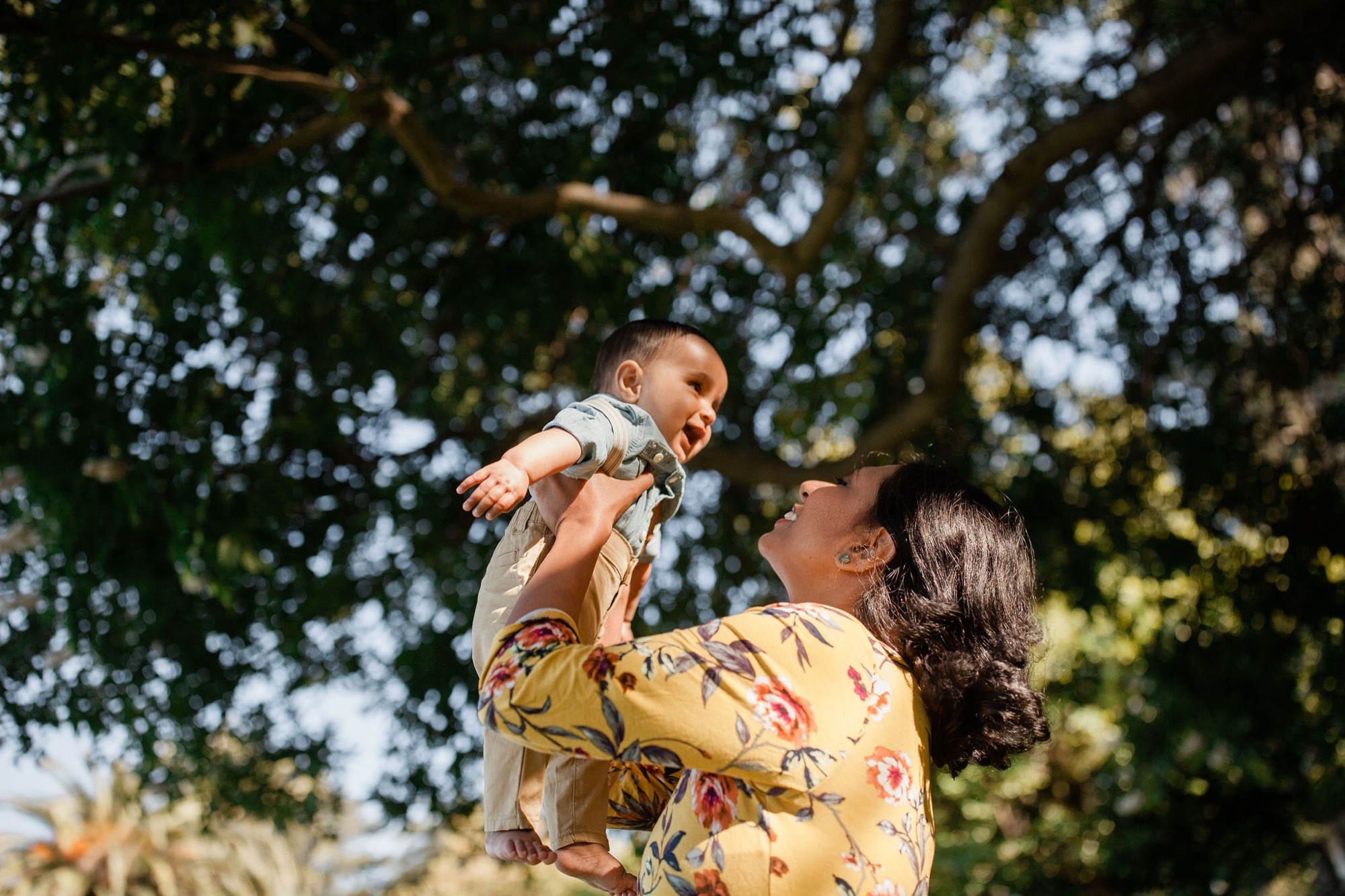 Mother lifts her laughing toddler toward dappled sunlight filtering through oak tree branches, his arms stretched wide in pure joy